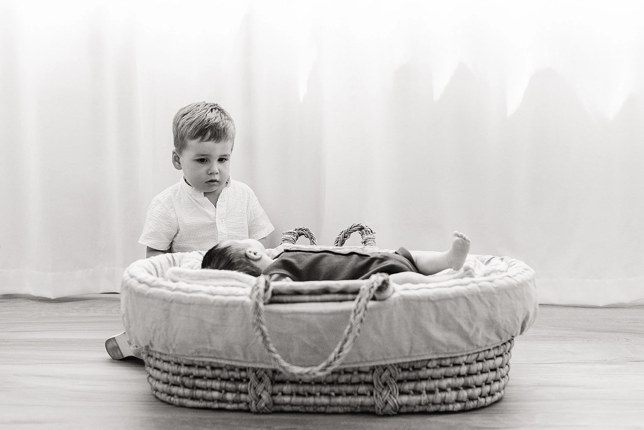A young boy looking at a baby lying in a woven basket on the floor during a studio lifestyle newborn session with Ally and B Photography in Naperville, IL.