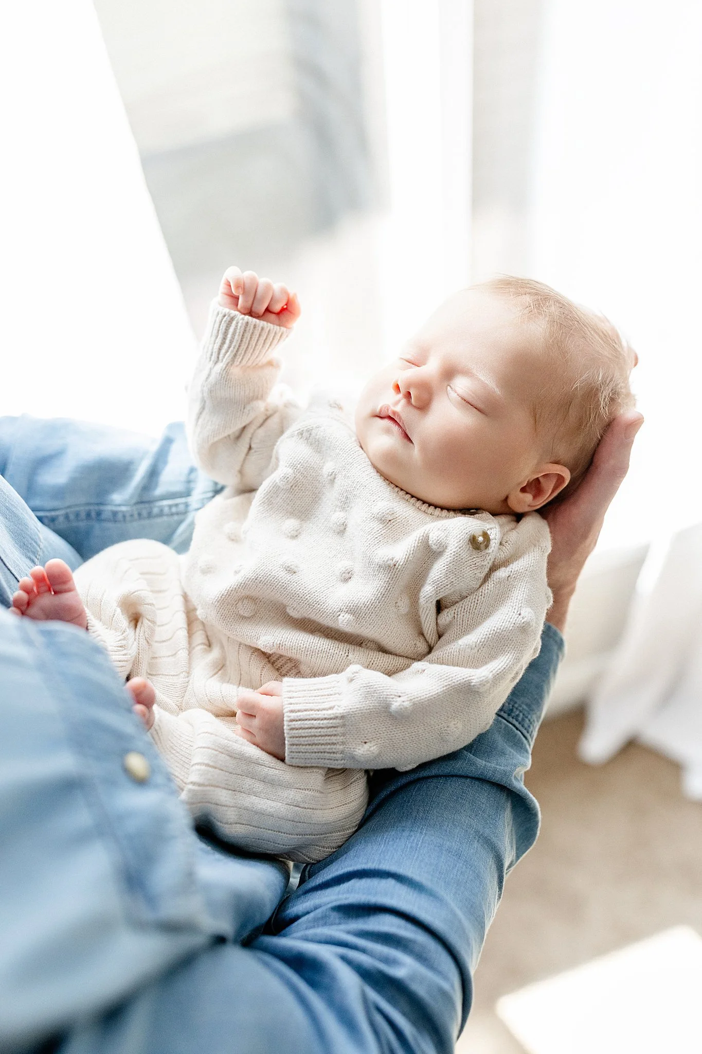 A sleeping baby with blonde hair and light skin, wearing a cream-colored sweater, resting in an adult's arms, in a bright room with sheer white curtains during newborn photos near me.