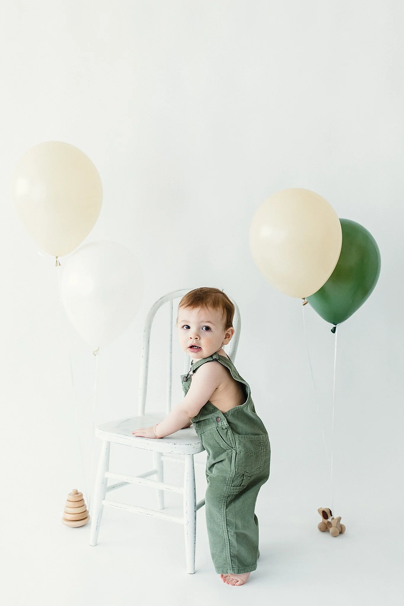 A young child in green overalls leaning on a white chair, surrounded by balloons and toys on a plain white background during milestone photos in Naperville.