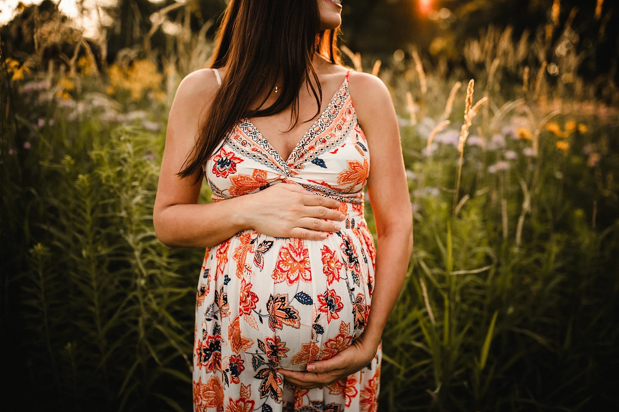A pregnant woman in a floral dress holding her belly in a field during sunset for Naperville maternity photos near me.