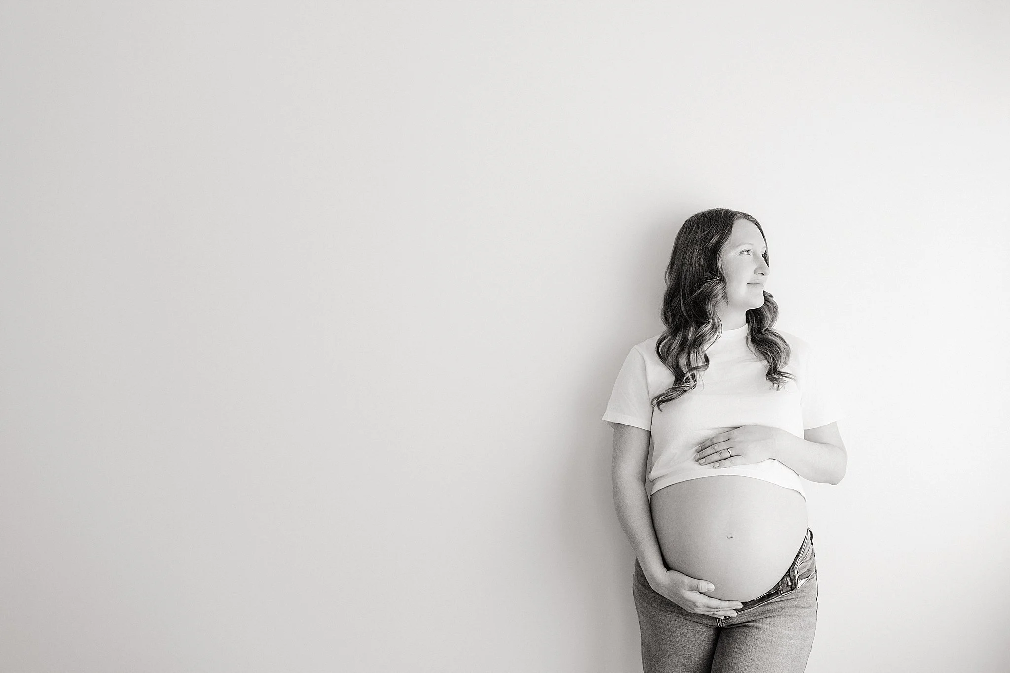 Black and white photo of a pregnant woman standing against a plain wall, smiling gently while holding her belly with one hand, and looking off to the side during a simple studio maternity session in downtown Naperville, IL.