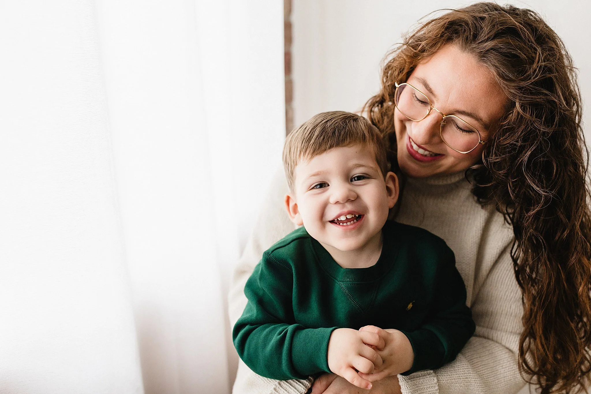 A woman with curly brown hair and glasses smiling while holding a young boy with short brown hair, both showing joyful expressions, indoors near a white curtain during a Naperville family photo session.