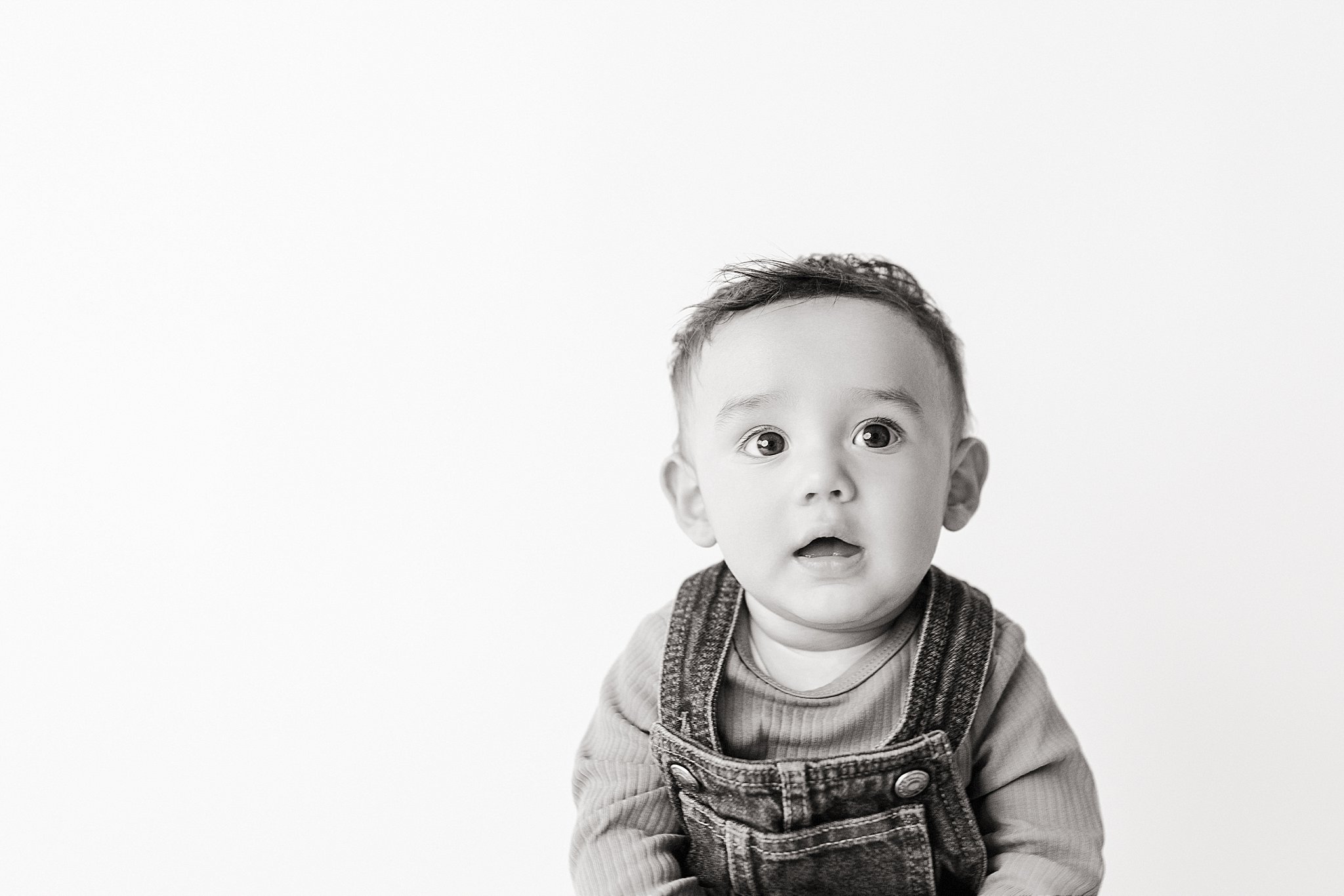 Black and white photo of a surprised baby boy with wide eyes and an open mouth, wearing overalls and a long sleeve shirt, against a plain white background during personality portraits in Naperville.