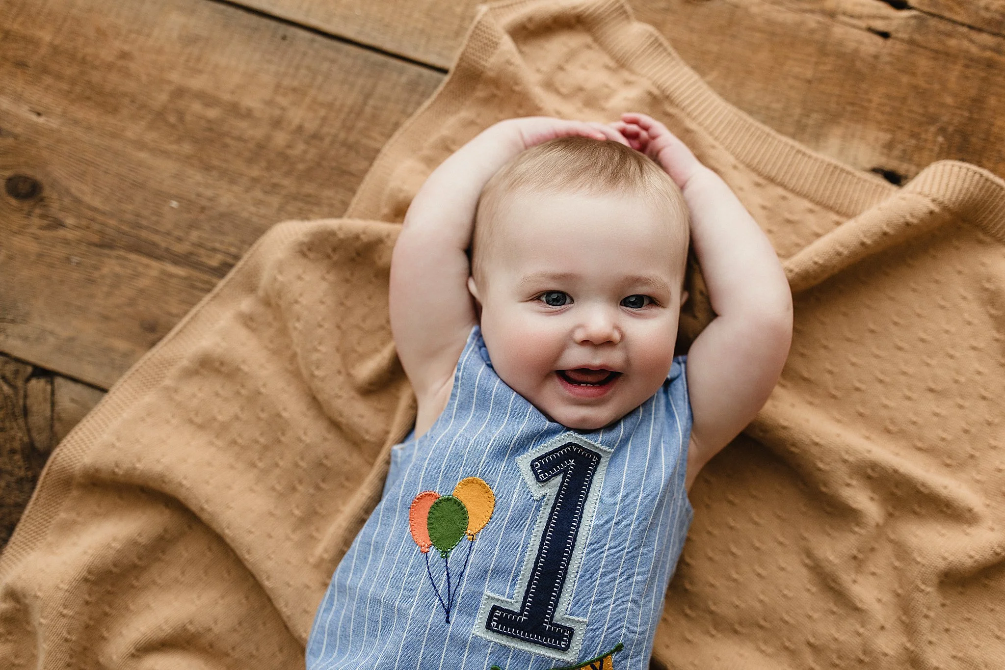 Smiling baby boy with blue eyes, lying on a beige blanket on a wooden floor, celebrating his first birthday, wearing a blue striped outfit with embroidered balloons and the number one during a baby photoshoot in Naperville.
