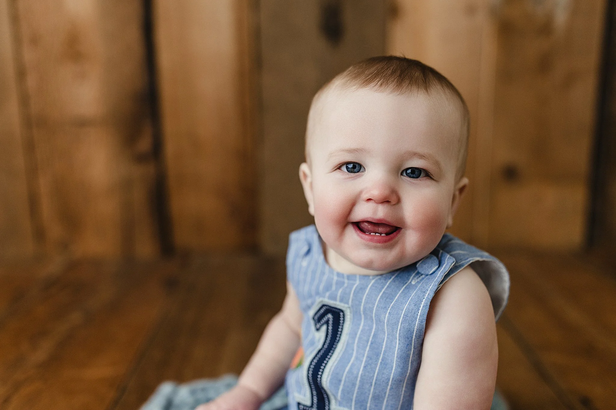 A smiling baby with blue eyes and short hair, wearing a sleeveless blue and white striped shirt, sitting on a wooden floor with a wooden wall in the background for first birthday photos near me.
