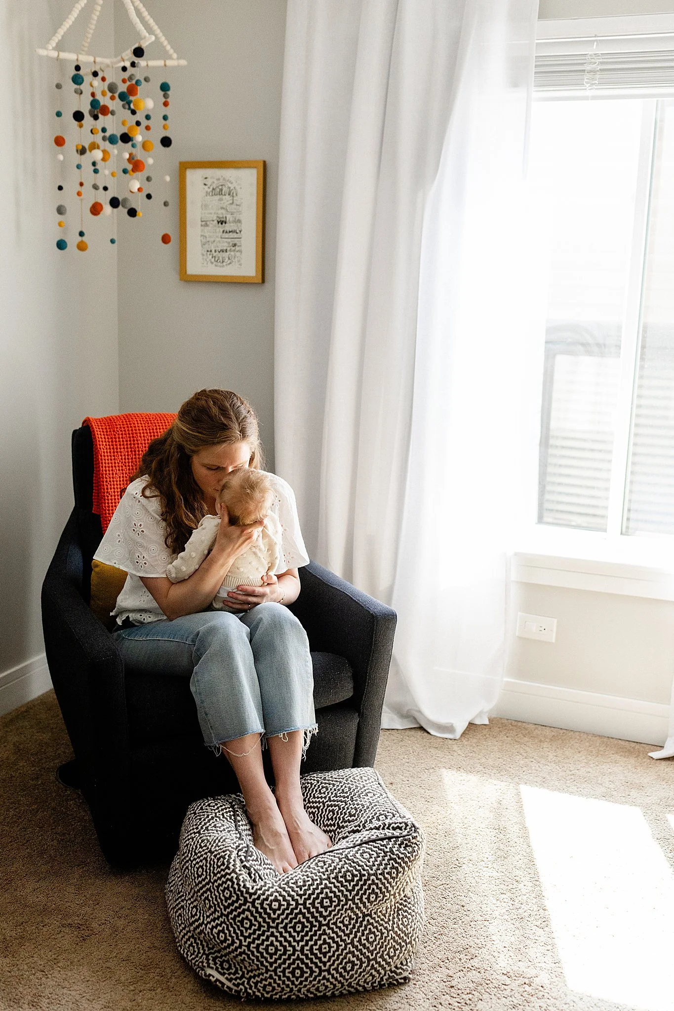 A woman sitting in a black armchair holding a small baby, with her feet resting on a patterned ottoman. They are in a bright room with white curtains and a window with Naperville lifestyle photography.