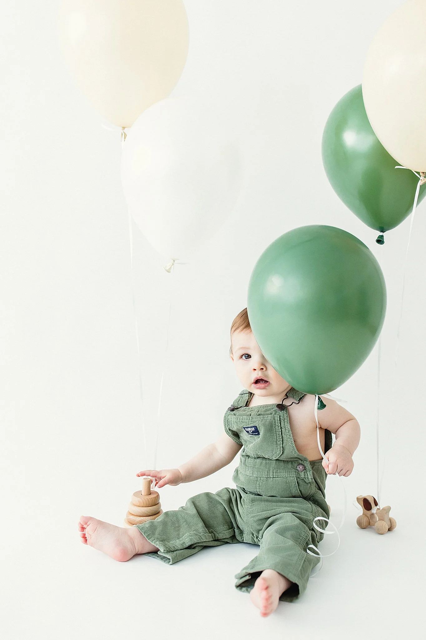 A baby sitting on the floor surrounded by green and white balloons, holding a wooden stacking toy, with a small wooden dog toy beside him for a one year old photoshoot with Ally and B Photography.