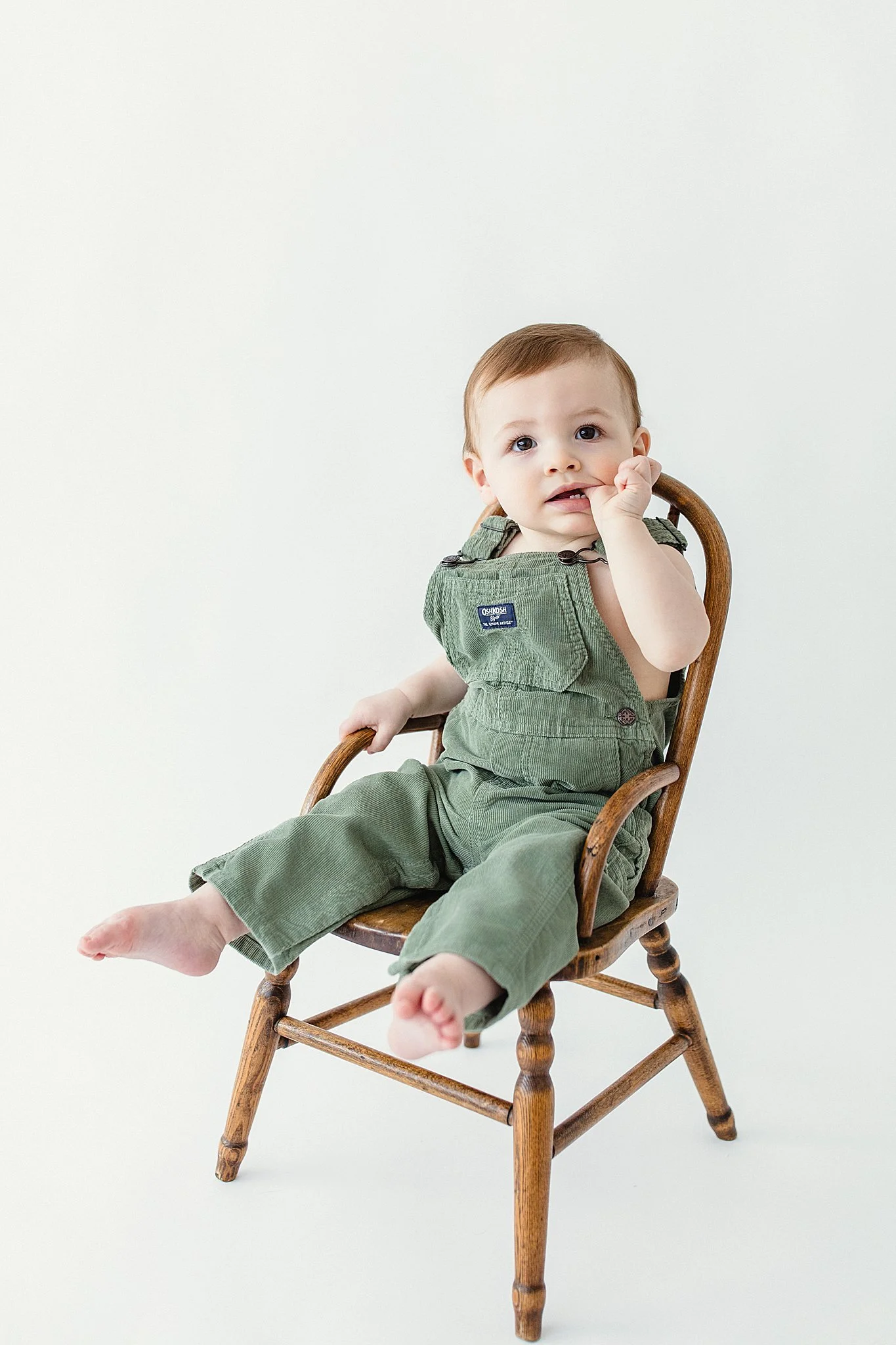 A young child with brown hair sitting on a wooden rocking chair against a plain white background, wearing green overalls and looking at the camera during a one year old session with Ally and B Photography.