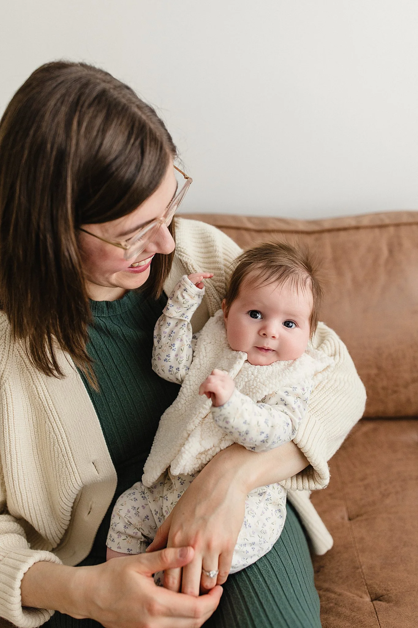 A woman with brown hair and glasses smiling while holding a baby girl with brown hair and big eyes sitting on her lap on a brown couch during family photos near me.