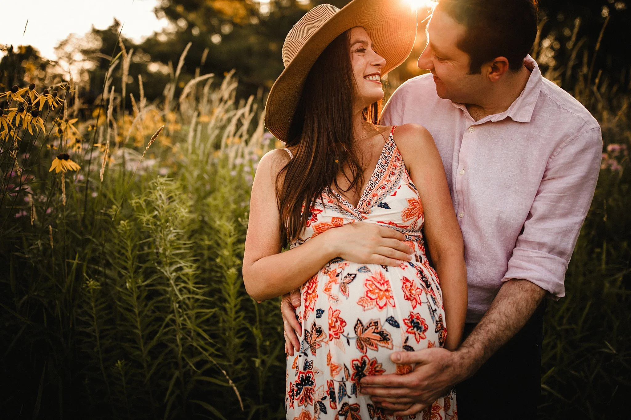 A pregnant woman and man in a field during sunset, smiling and looking at each other. The woman is wearing a floral dress and a sun hat, and the man is wearing a light pink shirt during a golden hour Naperville maternity photo session.