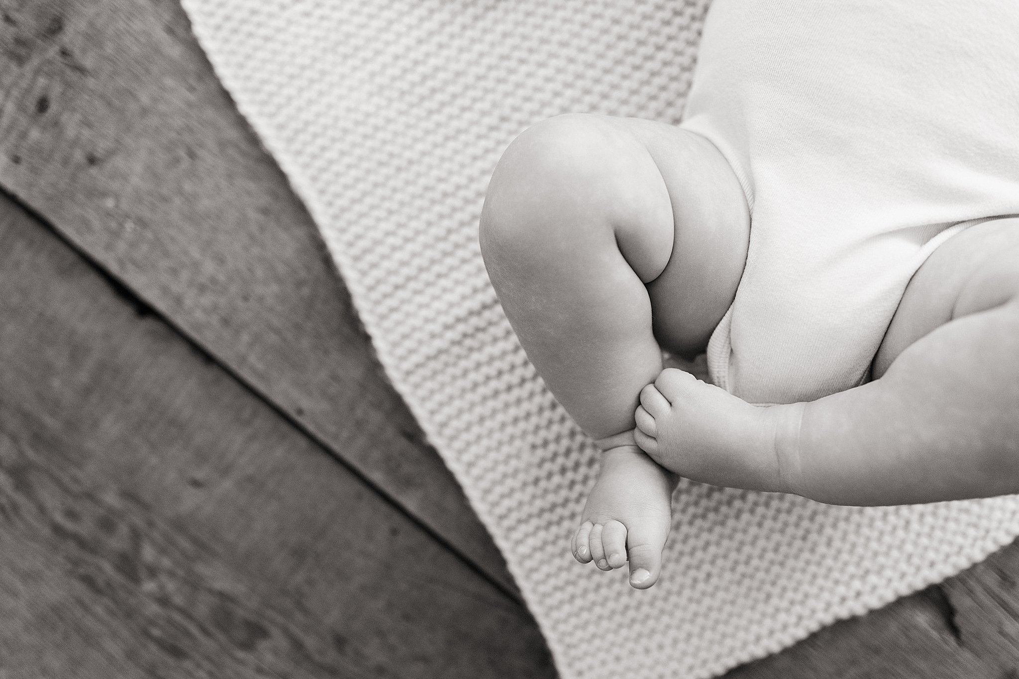 Black-and-white photo of a baby's legs and feet resting on a textured blanket with a wooden floor nearby during baby photos near me.