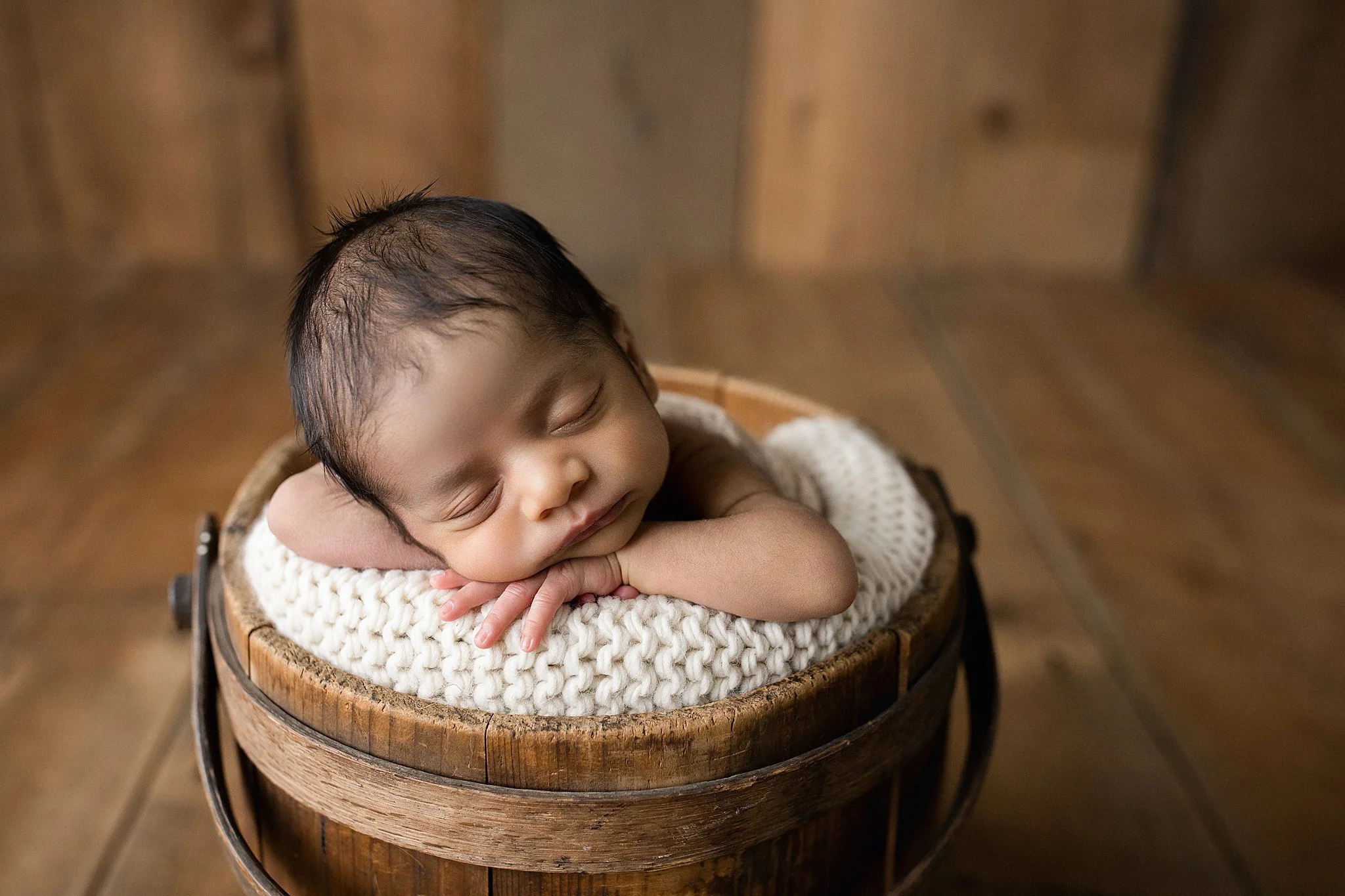 A sleeping baby with dark hair inside a wooden bucket lined with a white knitted blanket, resting their head on their arms, in a warm wooden room during a posed newborn session.