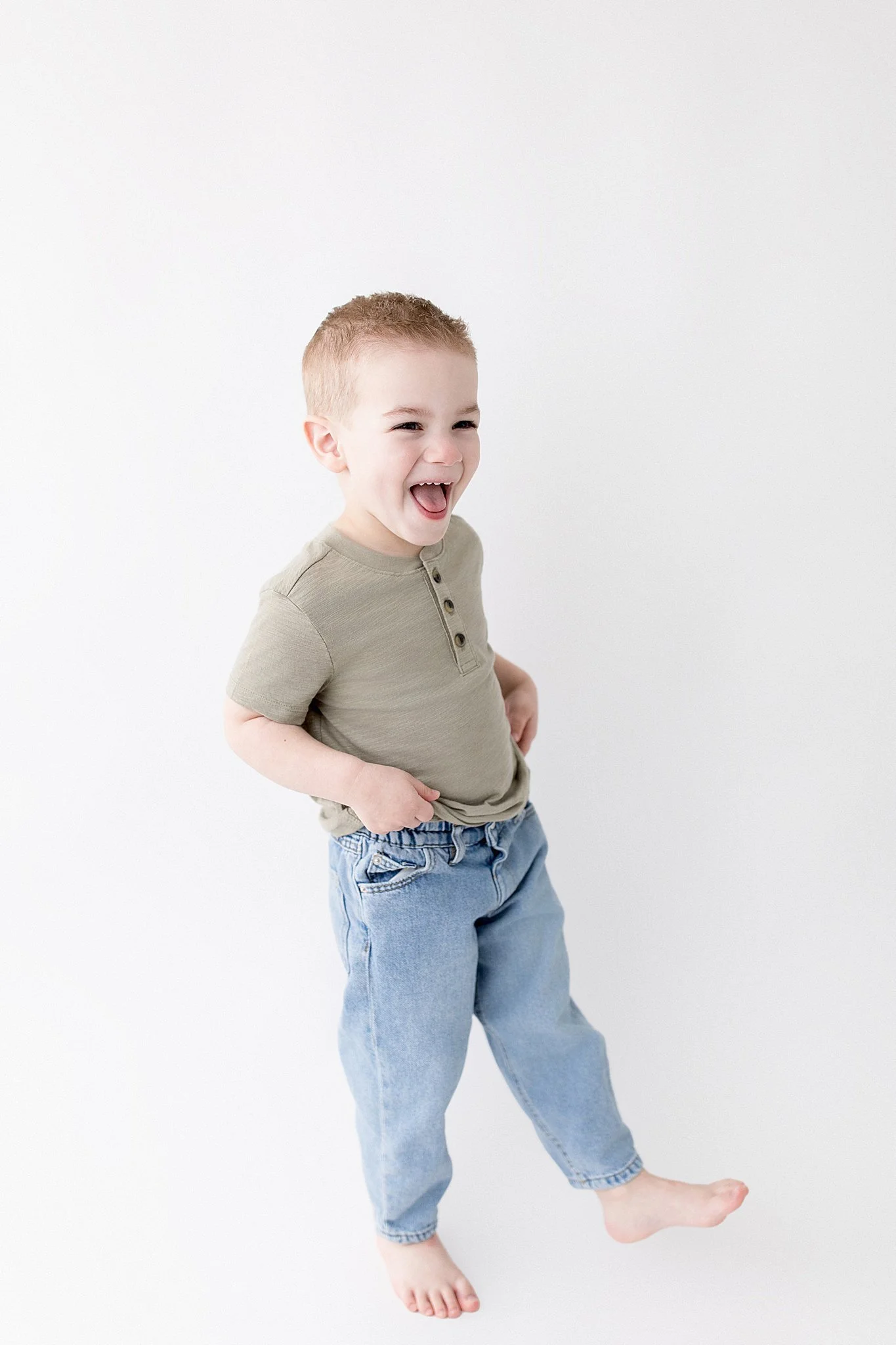 A young boy with short brown hair smiling and laughing, wearing a khaki t-shirt and baggy light-wash jeans, standing barefoot against a plain white background for photo studios in Naperville.