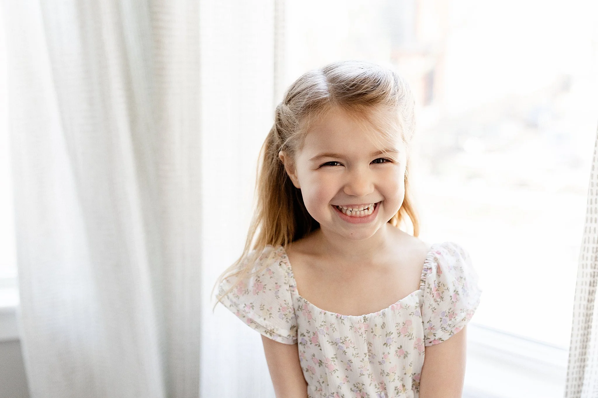 A young girl with blonde hair smiling and wearing a floral dress standing in front of a white curtain with sunlight shining through with a lifestyle photographer Aurora.