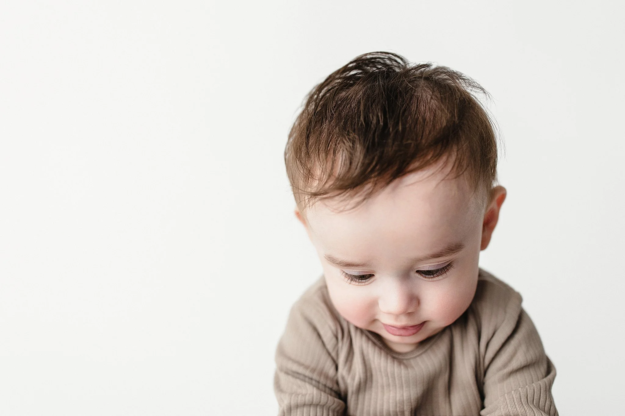 Close-up of a young boy with brown hair and fair skin, wearing a beige knit sweater, looking downward with a gentle expression, against a plain white background during a baby photoshoot Naperville.