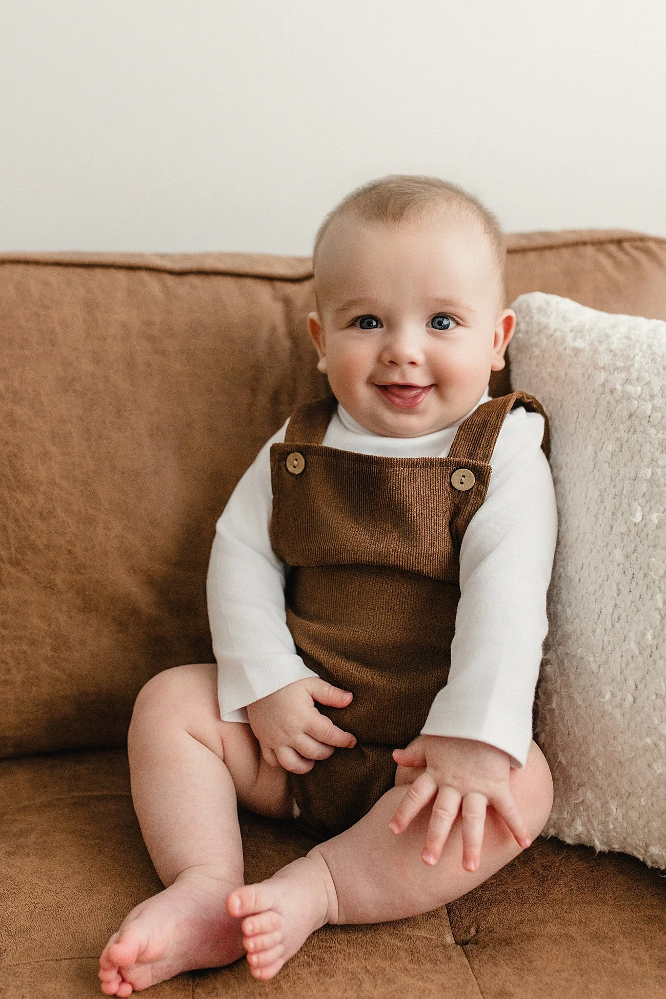 A smiling baby sitting on a brown sofa, wearing a white long-sleeve shirt and brown overalls, with a cream-colored pillow behind him for a baby lifestyle photoshoot near me.
