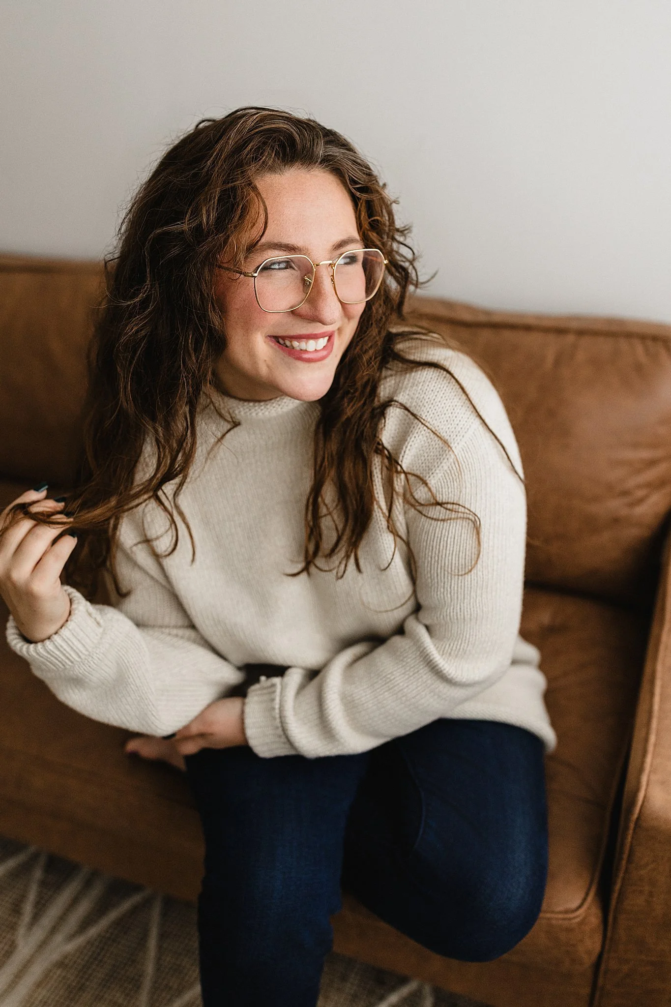 A smiling woman with curly brown hair and glasses wearing a cream sweater, sitting on a brown couch for personal brand photos in Naperville, IL.