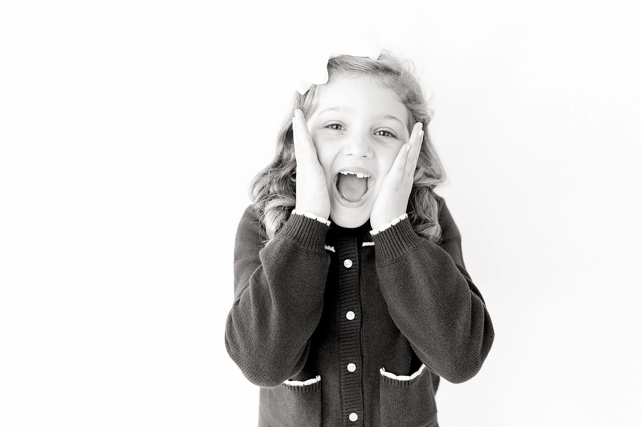 Black and white photo of a young girl with curly hair, wearing a dark cardigan, holding her face with her hands, and expressing surprise or excitement during a personality mini session in Naperville, IL.