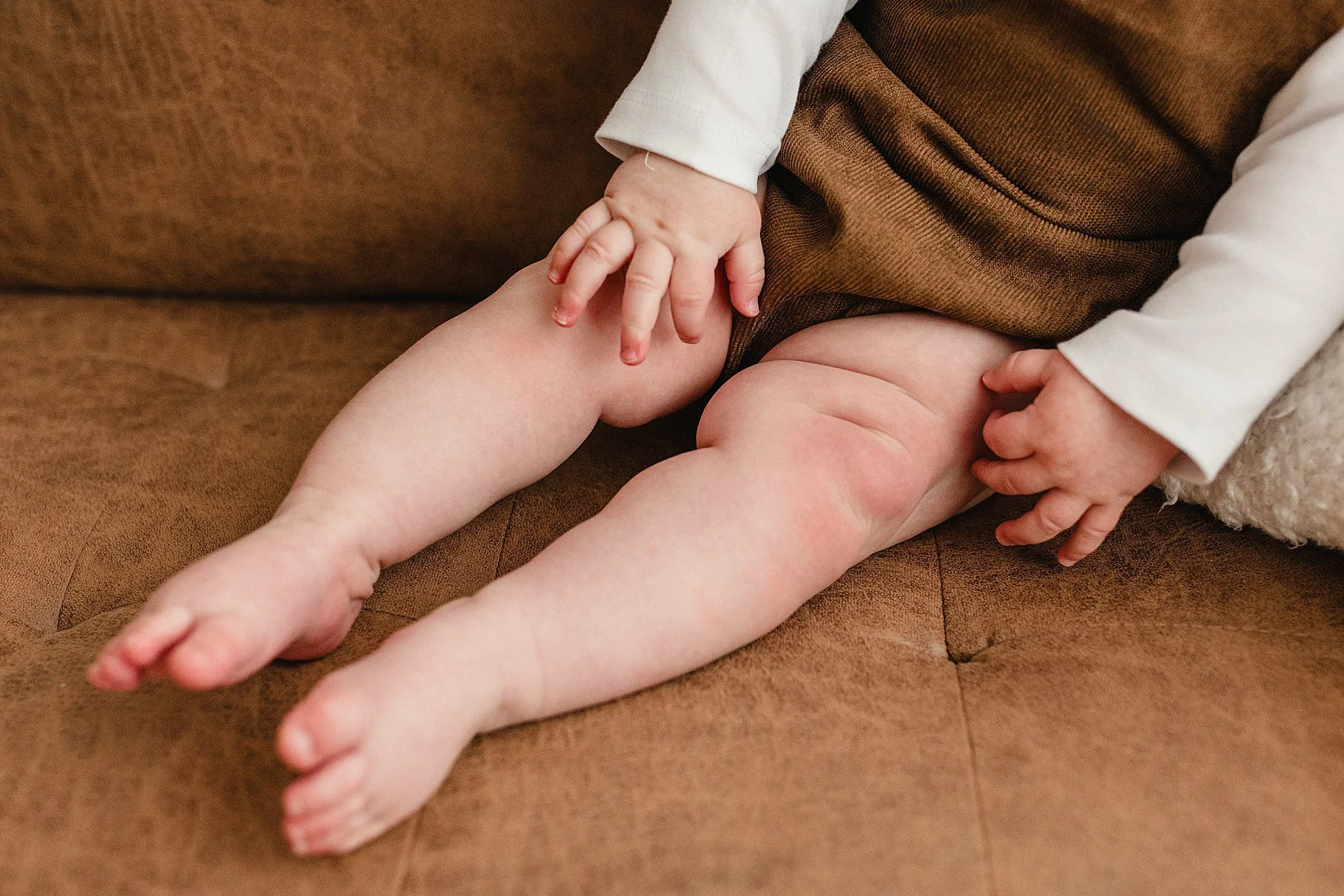 Close-up of a child's bare legs and hands resting on a brown couch, with one hand touching the knee and the other hand on the thigh for baby photos in downtown Naperville.