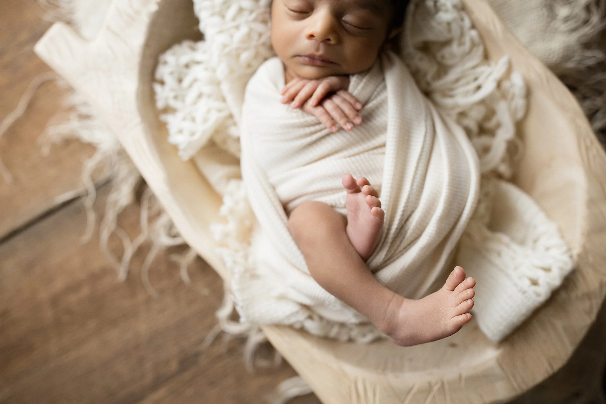 A sleeping baby wrapped in a beige blanket, lying on a soft blanket on a wooden surface with the best newborn photographer near me.