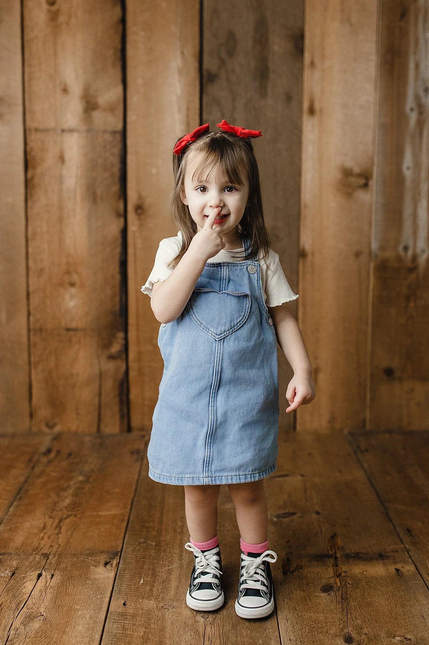 A young girl with brown hair, wearing a cream t-shirt, a denim dress, pink socks, and black-and-white sneakers, standing against a wooden wall, touching her nose with her finger during milestone photos for kids.