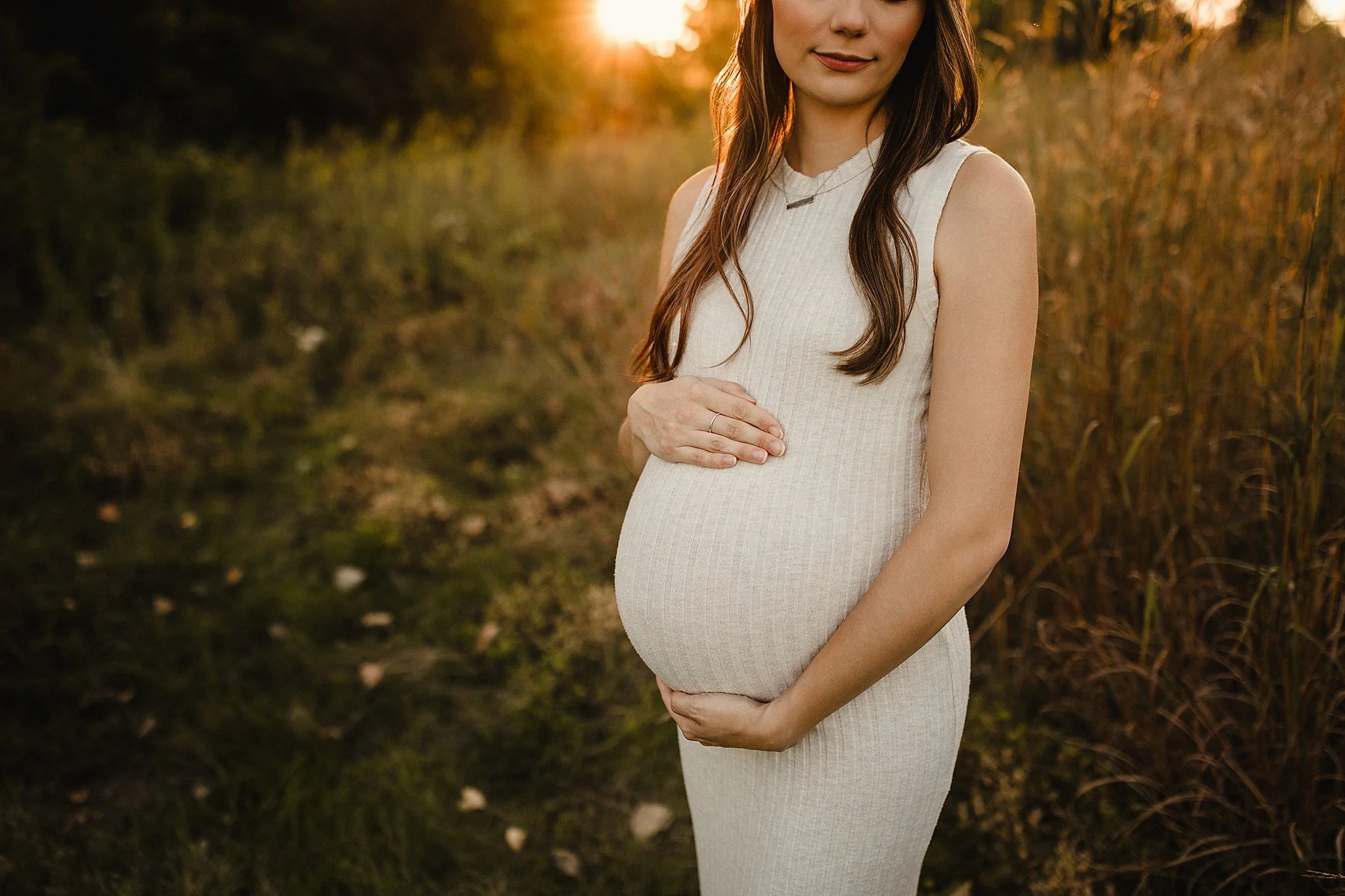 A pregnant woman stands outdoors holding her belly with both hands, dressed in a sleeveless cream-colored dress, with long brown hair, during sunset in a natural setting during a Naperville maternity photography session.