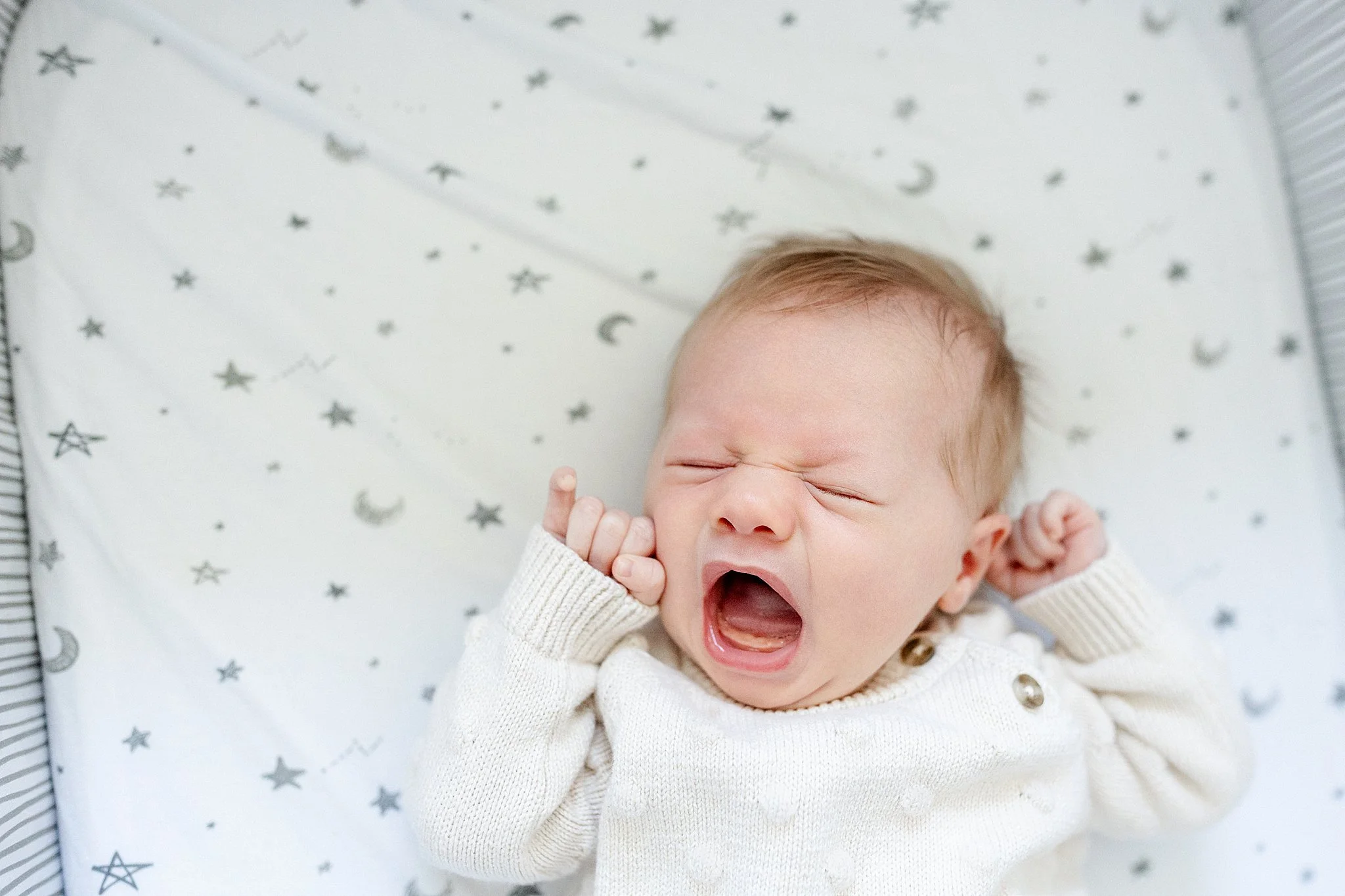 A toddler with light skin and blonde hair yawning, lying on a white blanket decorated with gray stars and moons during a lifestyle baby photoshoot in Naperville.