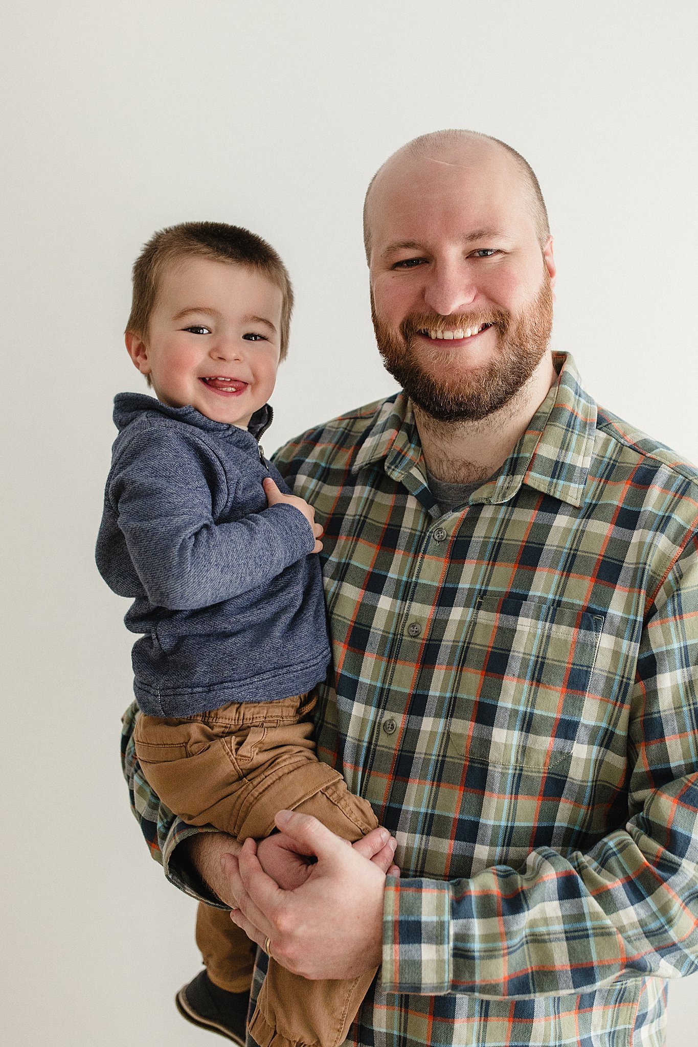 A smiling man with a beard holding a happy toddler boy with short hair, both looking at the camera, against a plain white background with Ally and B Photography in Naperville, IL.