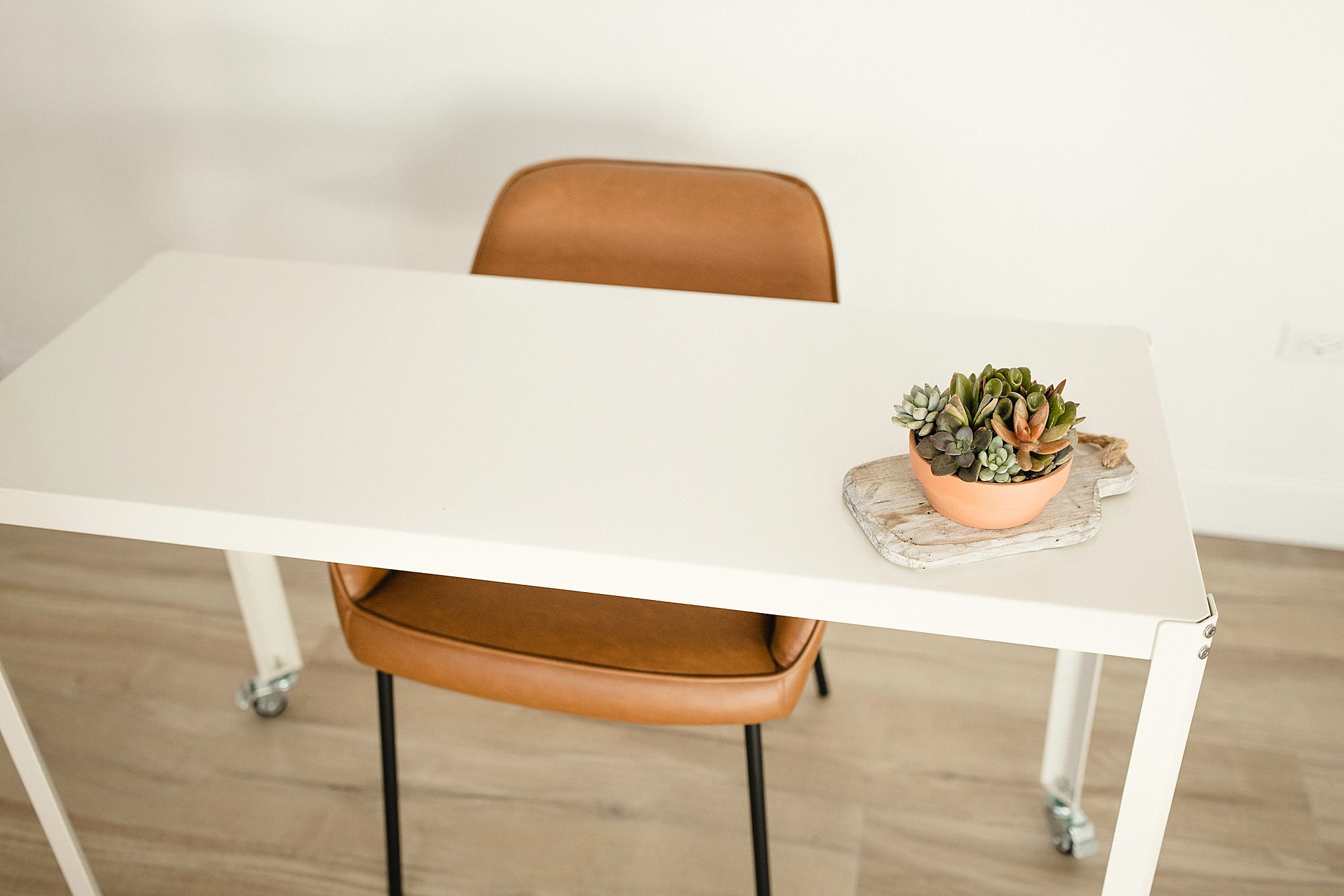 A white desk with a potted succulent plant on a wooden tray, a brown chair behind the desk, and light-colored wooden flooring in photography studios in Naperville for branding photos.