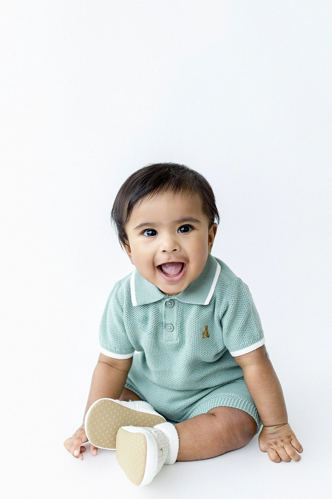 Smiling baby boy sitting on the floor in a light green outfit with white trim, holding his shoes, against a plain white background with baby photographers near me.