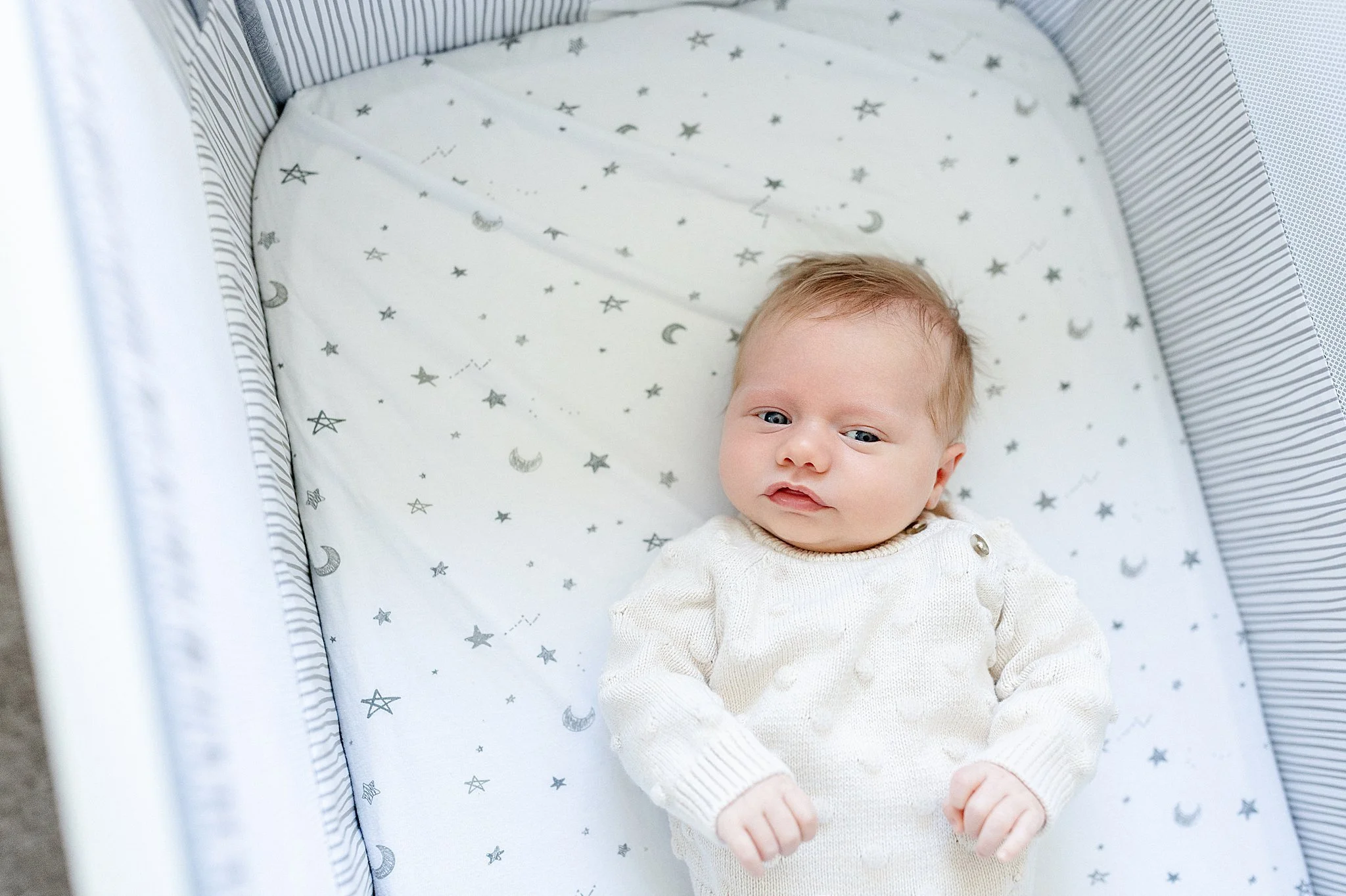 A baby lying in a crib with star and moon patterned bedding, wearing a cream-colored sweater with a lifestyle photographer Chicago.