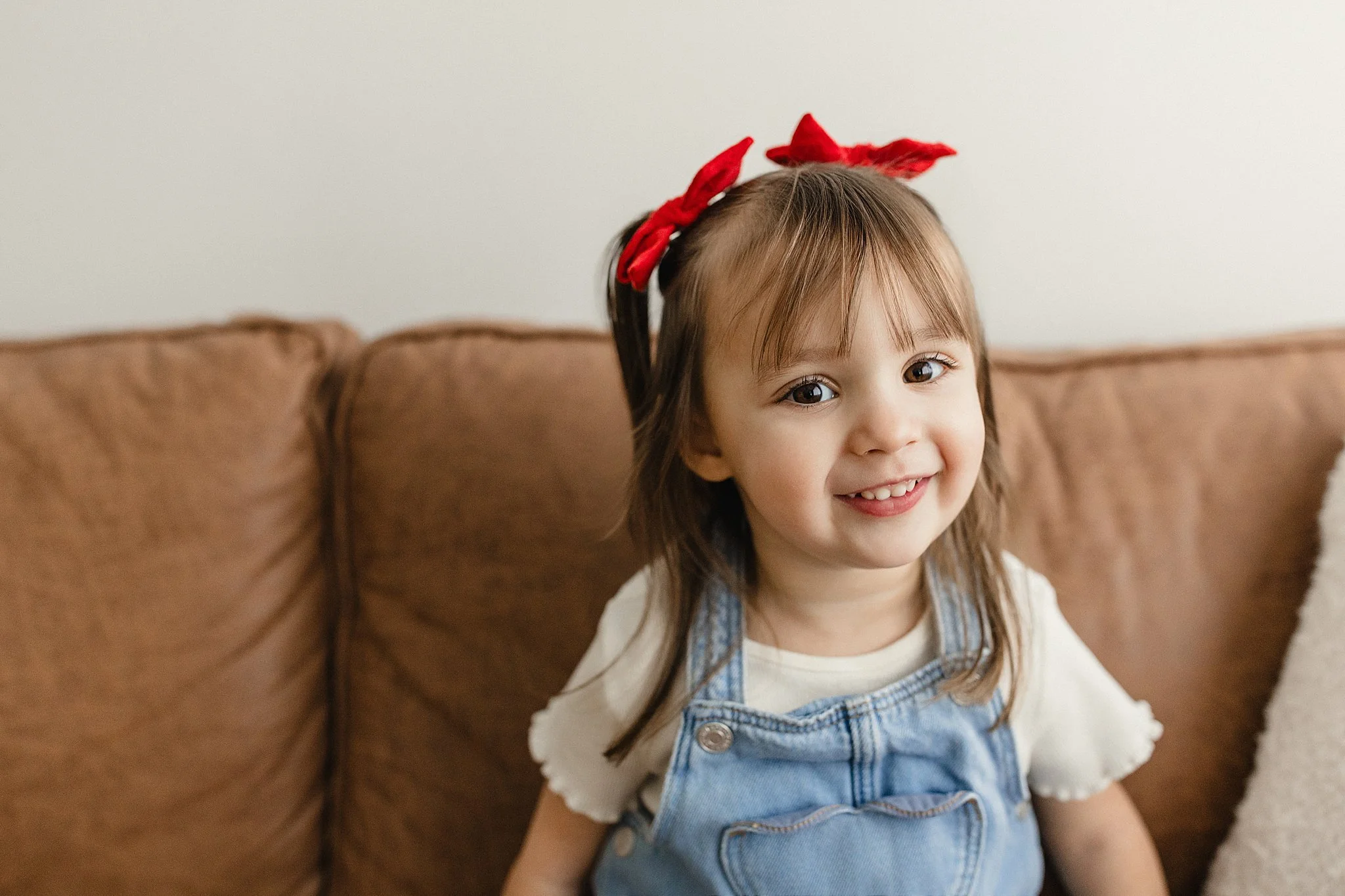 Smiling young girl with brown hair and red bow sitting on a beige couch for kids photography near me.