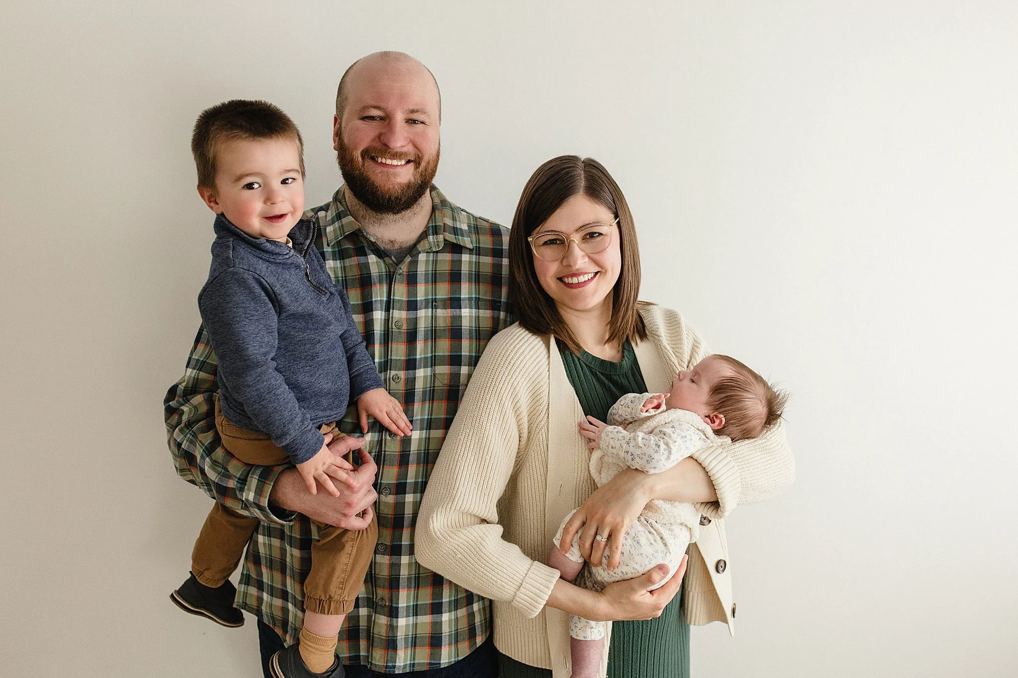 A smiling family of four, including a baby girl, stands together against a plain off-white wall. The mother holds the baby, while the father carries a young boy on his back. The family appears happy and relaxed during studio photos naperville.
