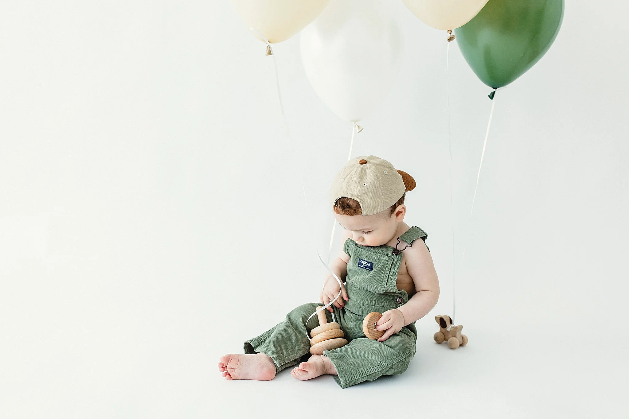 A young child sitting on a white background holding wooden toys, wearing green overalls, a beige cap, and looking down at toys during a one year photoshoot in Naperville, IL.
