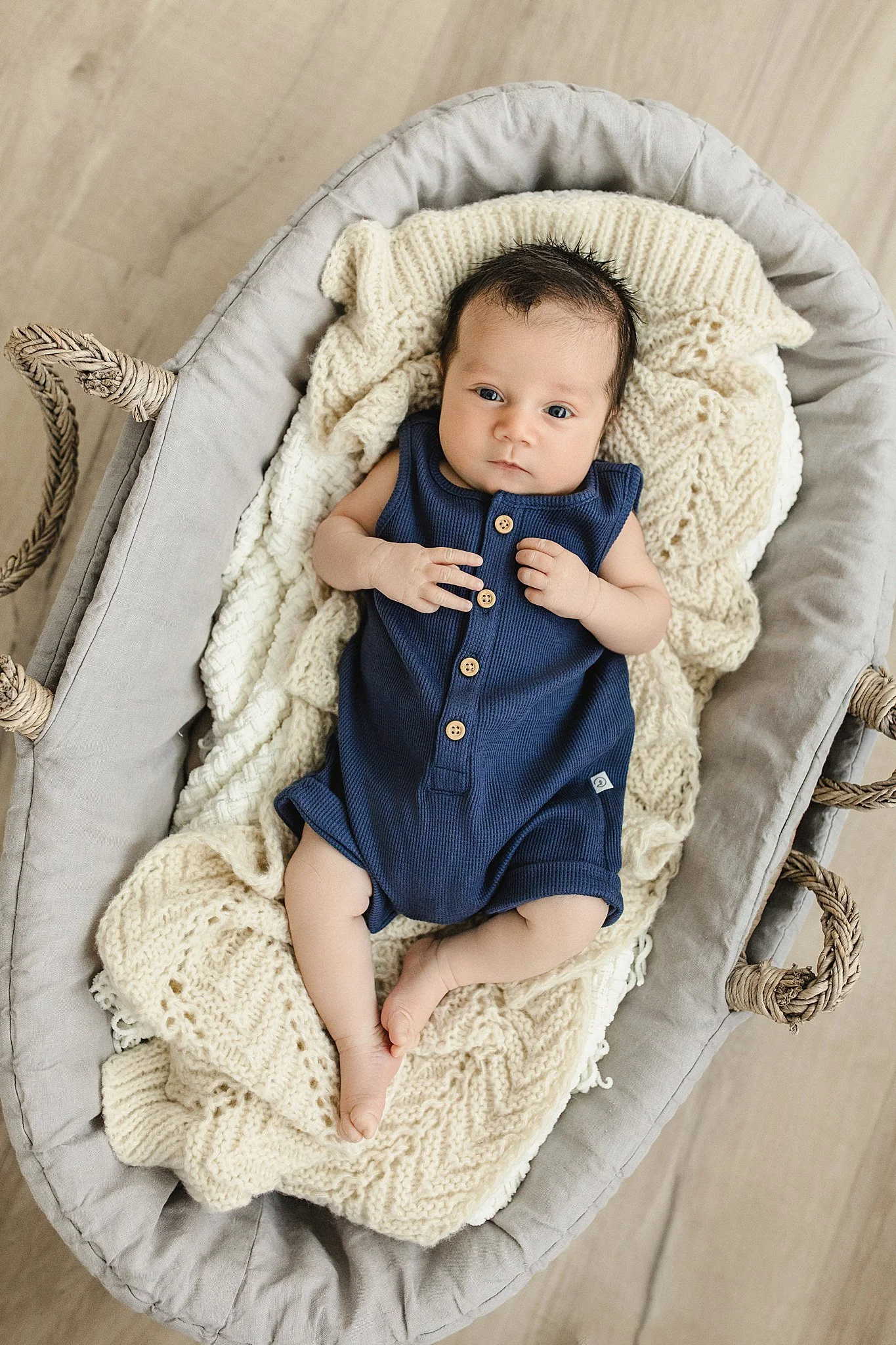 A baby with dark hair and blue eyes lying in a cozy gray basket lined with a chunky cream blanket, wearing a navy blue sleeveless romper with buttons during lifestyle newborn photos Naperville.