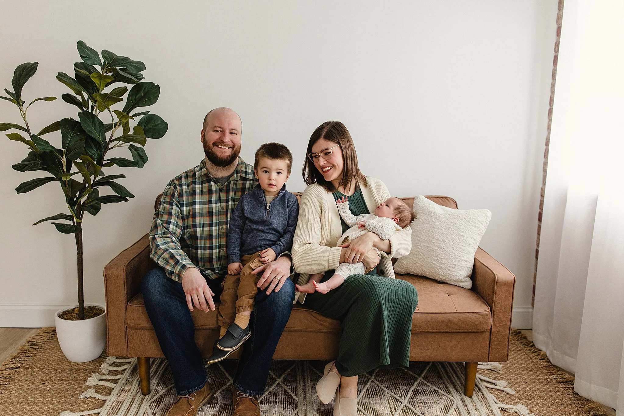 A happy family of four sitting on a brown sofa in a bright living room. The father, mother, young boy, and baby girl are smiling and enjoying their time together during lifestyle family photos near me.