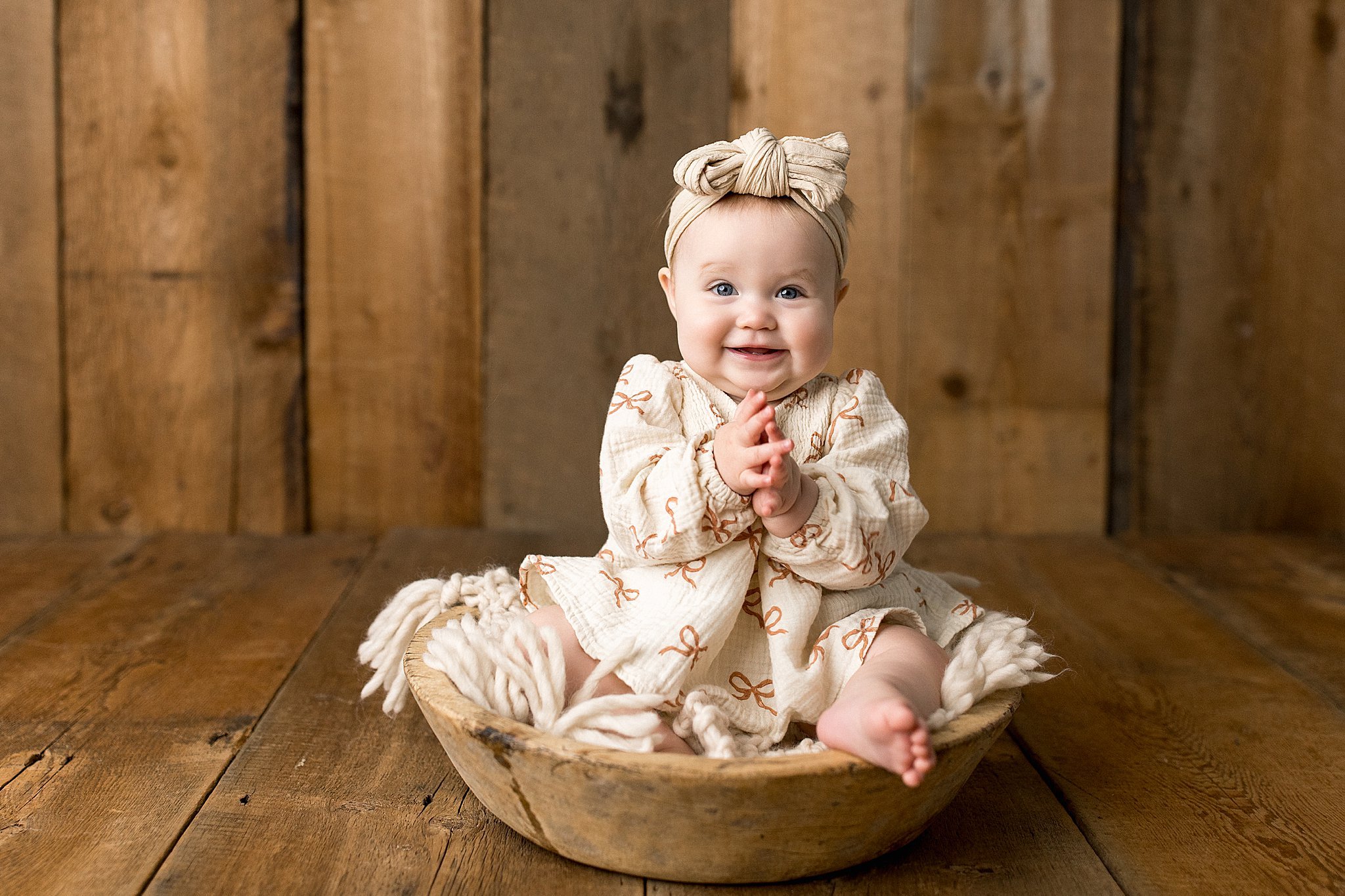 A smiling baby girl with blue eyes and a beige bow headband, sitting in a wooden bowl on a wooden floor against a wooden backdrop, wearing a beige dress with brown bow patterns, with her hands clasped together for cute baby photoshoot near me.
