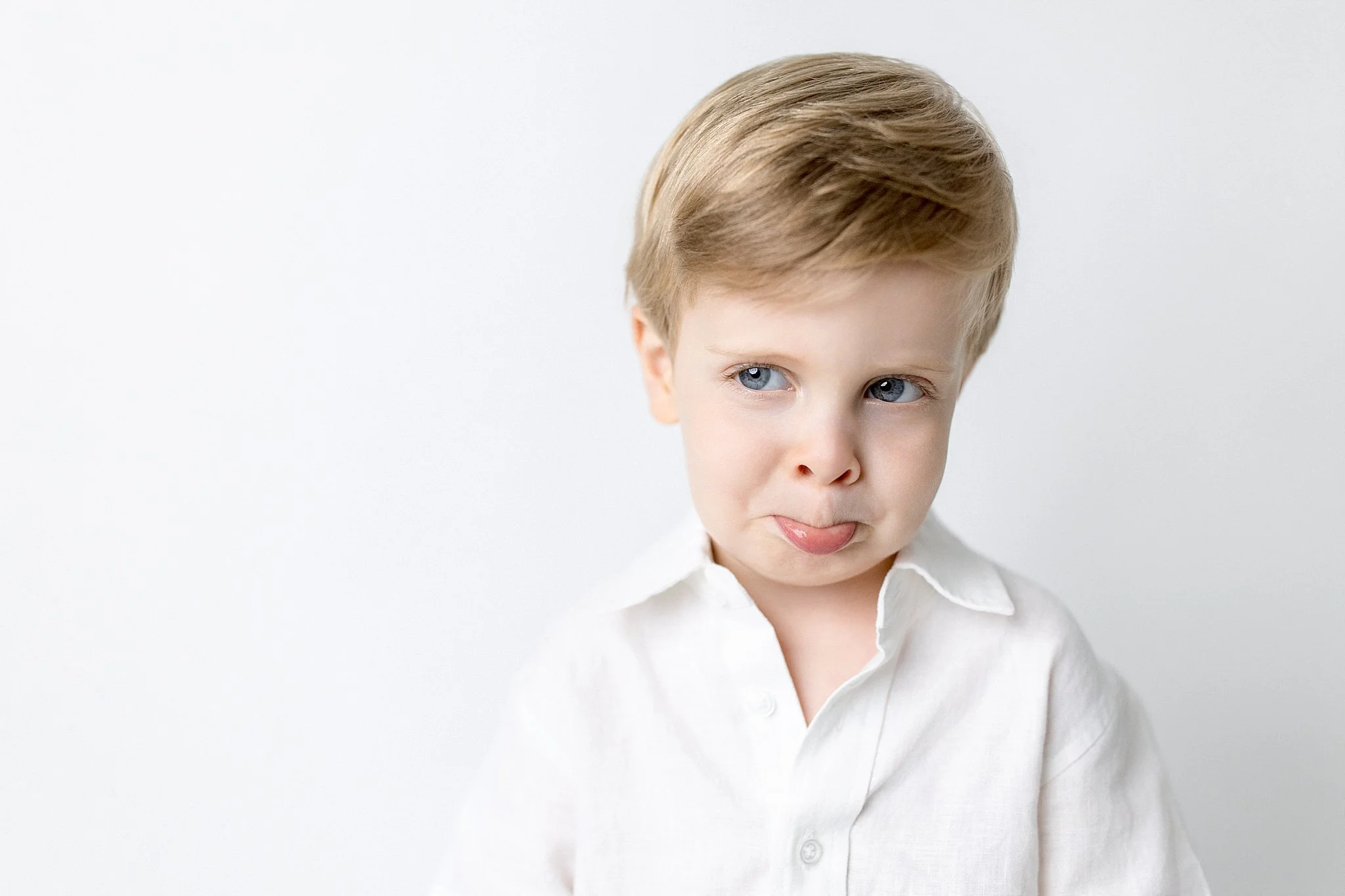 A young boy with blonde hair and blue eyes, wearing a white shirt, making a playful expression with his tongue sticking out slightly, against a plain light background for a personality portrait session in Naperville.