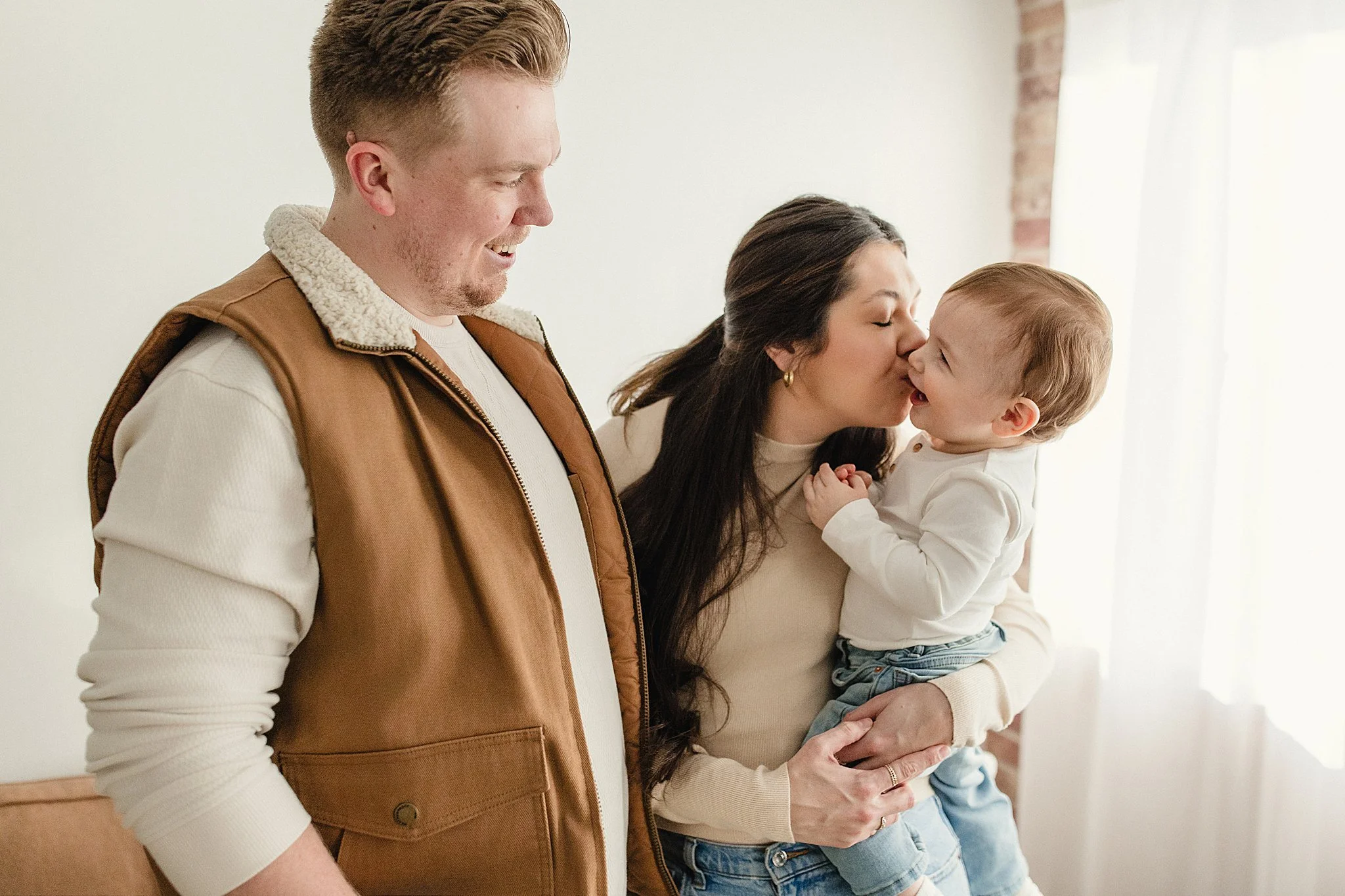 A woman holding a young boy while kissing him on the cheek, with a man standing nearby smiling with the best family photographers in Naperville, IL.