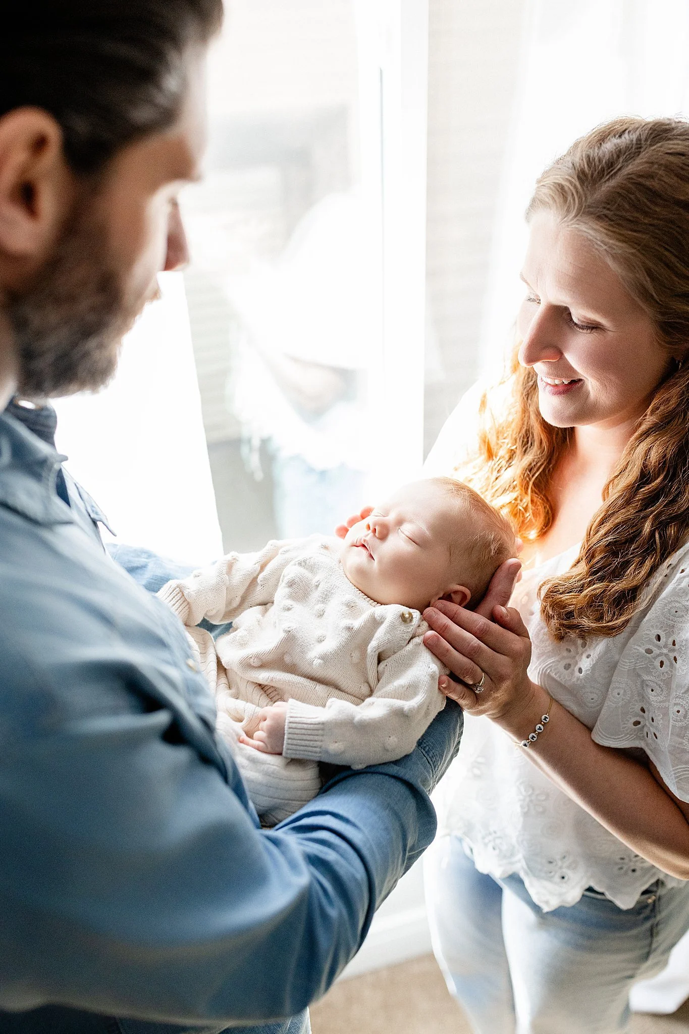 A woman with red hair smiling and holding a sleeping baby, with a man with dark hair and beard looking on, in a bright room near a window for lifestyle photographer Naperville.