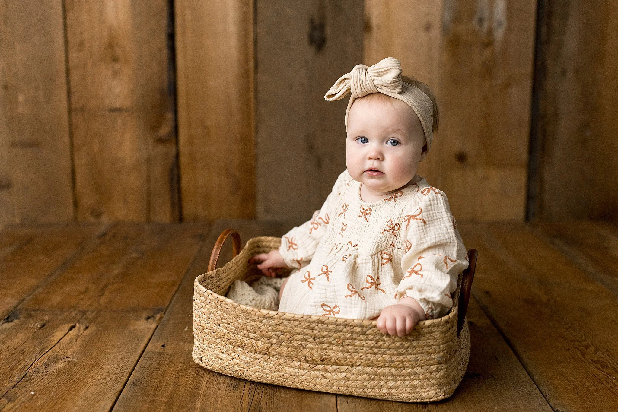 A baby girl with blue eyes sitting in a woven basket on a wooden floor, wearing a beige headband with a bow and a cream-colored dress with brown ribbon patterns. The background consists of wooden planks for a one year old photoshoot in Naperville, IL