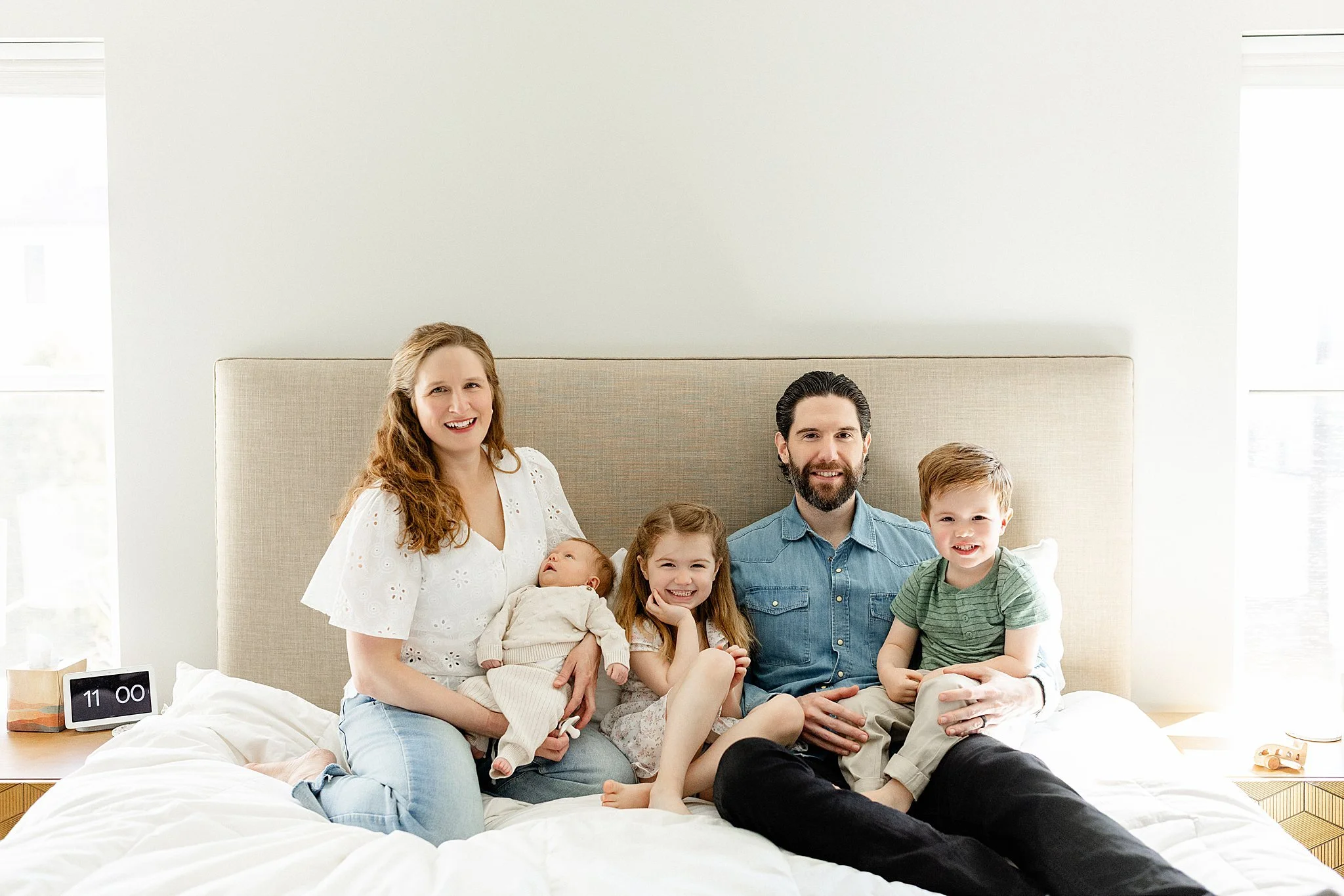 Family of seven sitting on a bed in a modern, bright bedroom. The group includes three children, two women, and two men. They are smiling and appear happy during an in home newborn session near me.
