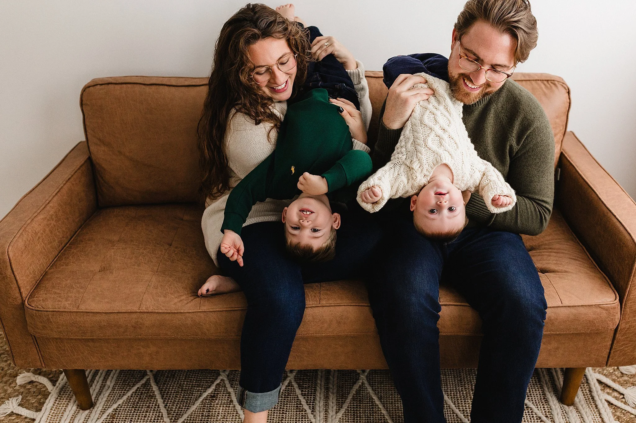 A smiling family of four, a man, woman, and two children, sitting on a brown sofa in a living room. The children are playful, with one upside down and the other hanging upside down from the man's lap during a studio family photoshoot near Naperville.