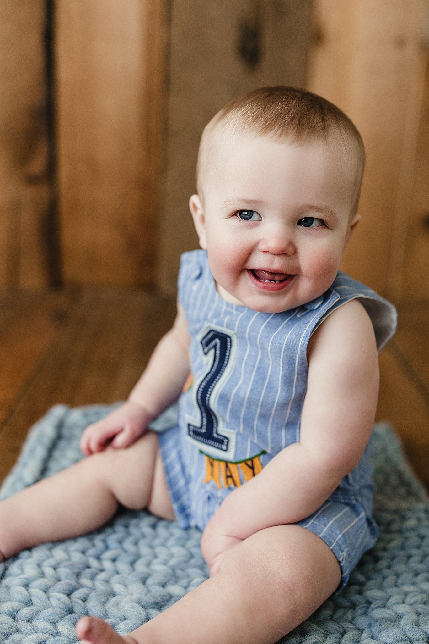 A smiling baby with blue eyes, wearing a blue striped outfit with the number 1 and the word 'HAPPY' on it, sitting on a textured gray blanket on a wooden floor with wooden wall in the background during a baby photoshoot in Naperville, IL.