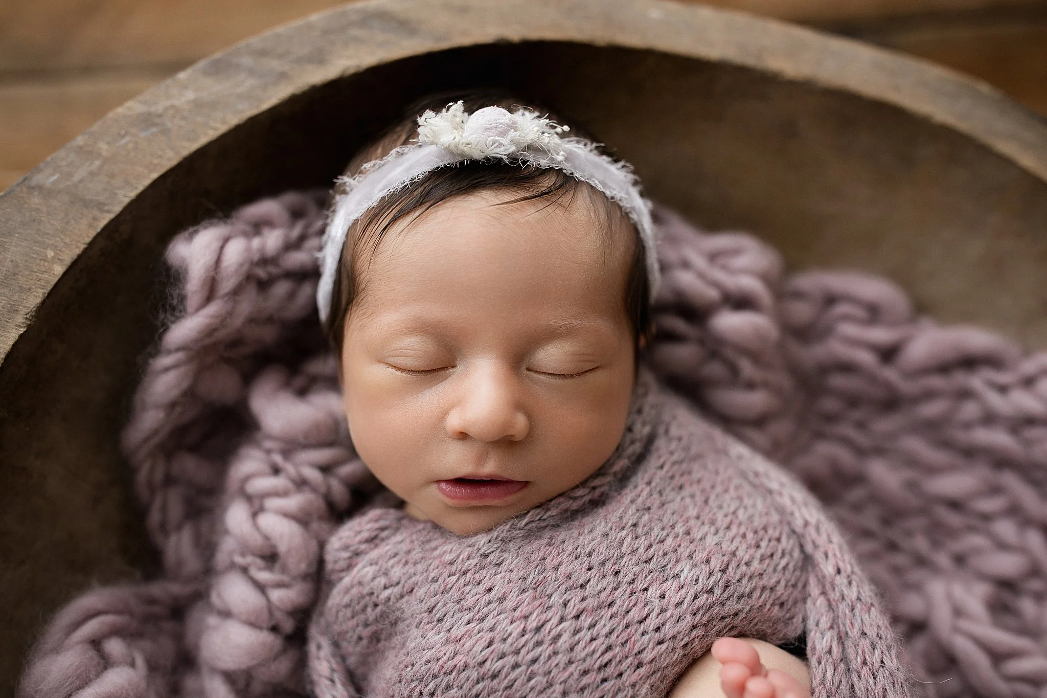 Close-up of a sleeping newborn baby girl with a white headband, wrapped in pink knit blankets, lying in a wooden bowl during posed newborn photography.