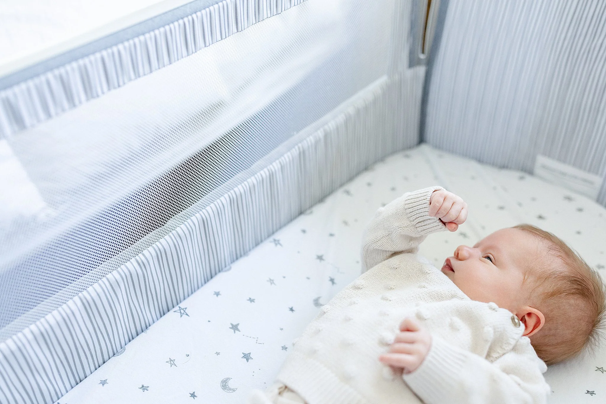 A baby lying on a bed with star and moon patterned sheets, dressed in a cream-colored sweater, reaching up with one hand, in a crib with light-colored vertical striped fabric and small circular perforations.