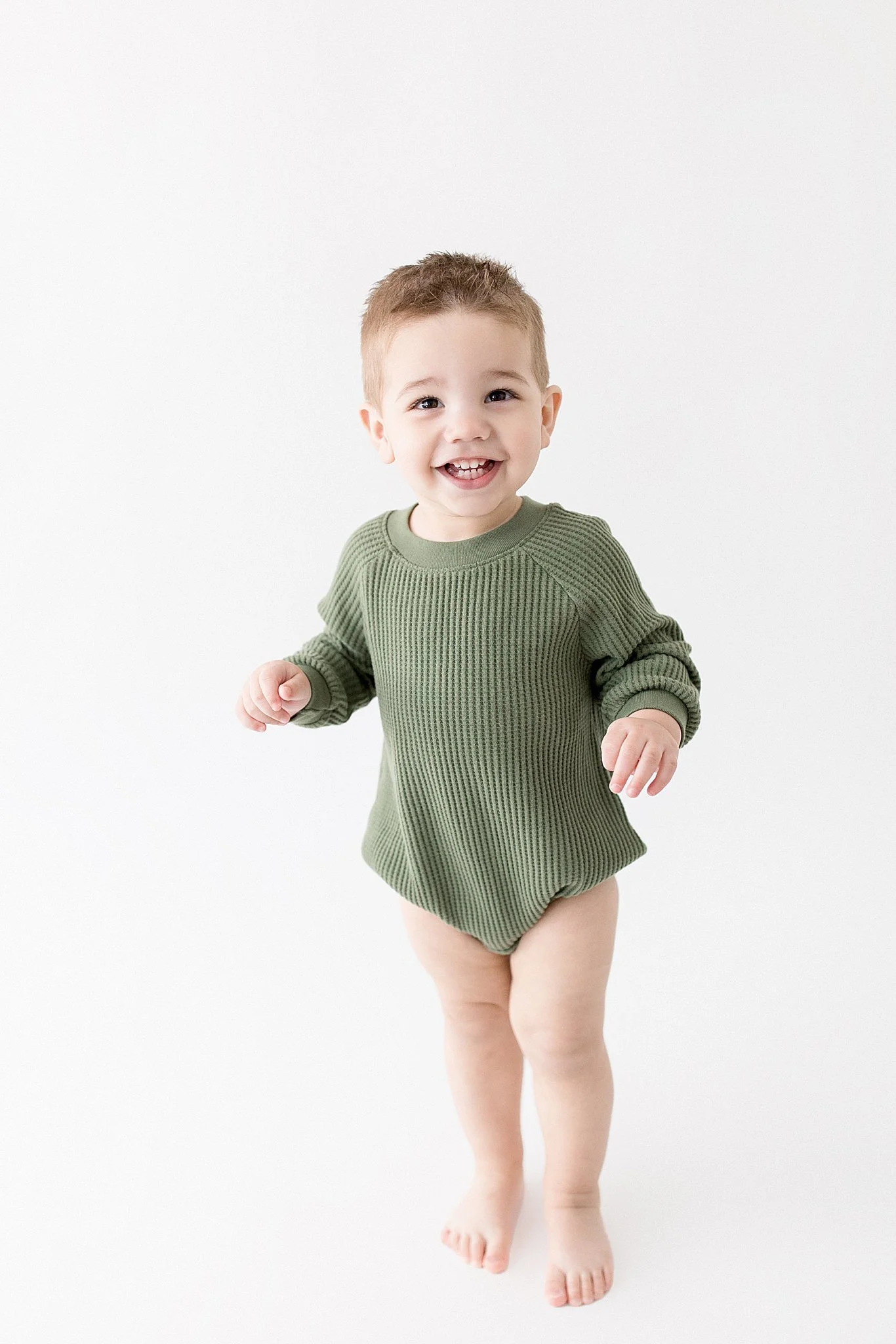 A young child with short brown hair, smiling, running barefoot in a green ribbed long sleeve shirt, against a plain white background for photo studios in downtown naperville, il.