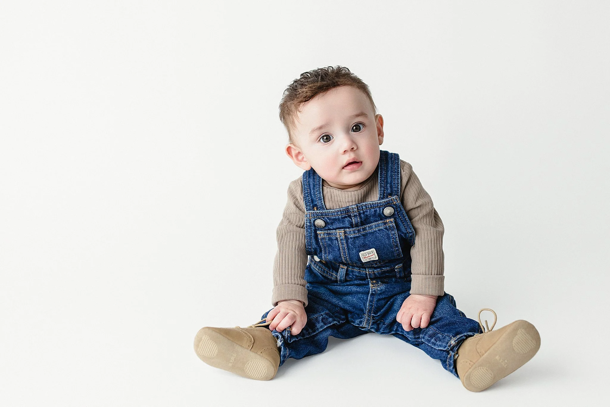 A young child sitting on a plain white background, wearing denim overalls, a beige long-sleeve shirt, and tan shoes, looking at the camera with a curious expression.