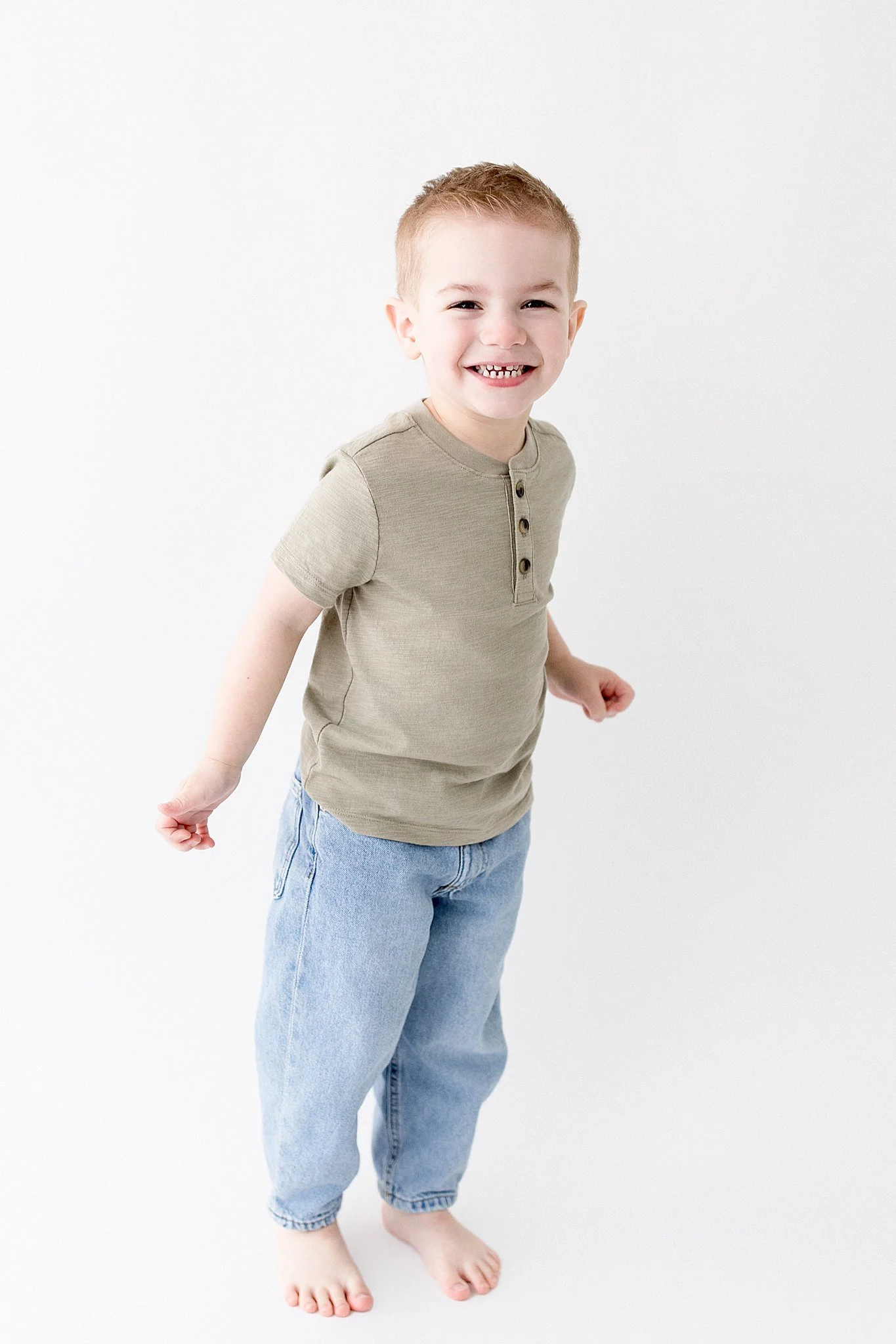 A smiling young boy with short red hair wearing a beige short-sleeve shirt and baggy light blue jeans standing on a plain white background with the best kids photographer in Naperville, IL.