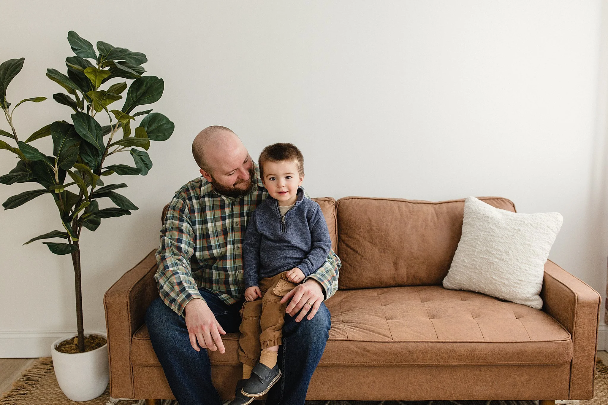 A man with a beard and a child sitting on a brown sofa in a living room, with a large green plant in a white pot beside them and a white pillow on the sofa during family photography naperville.